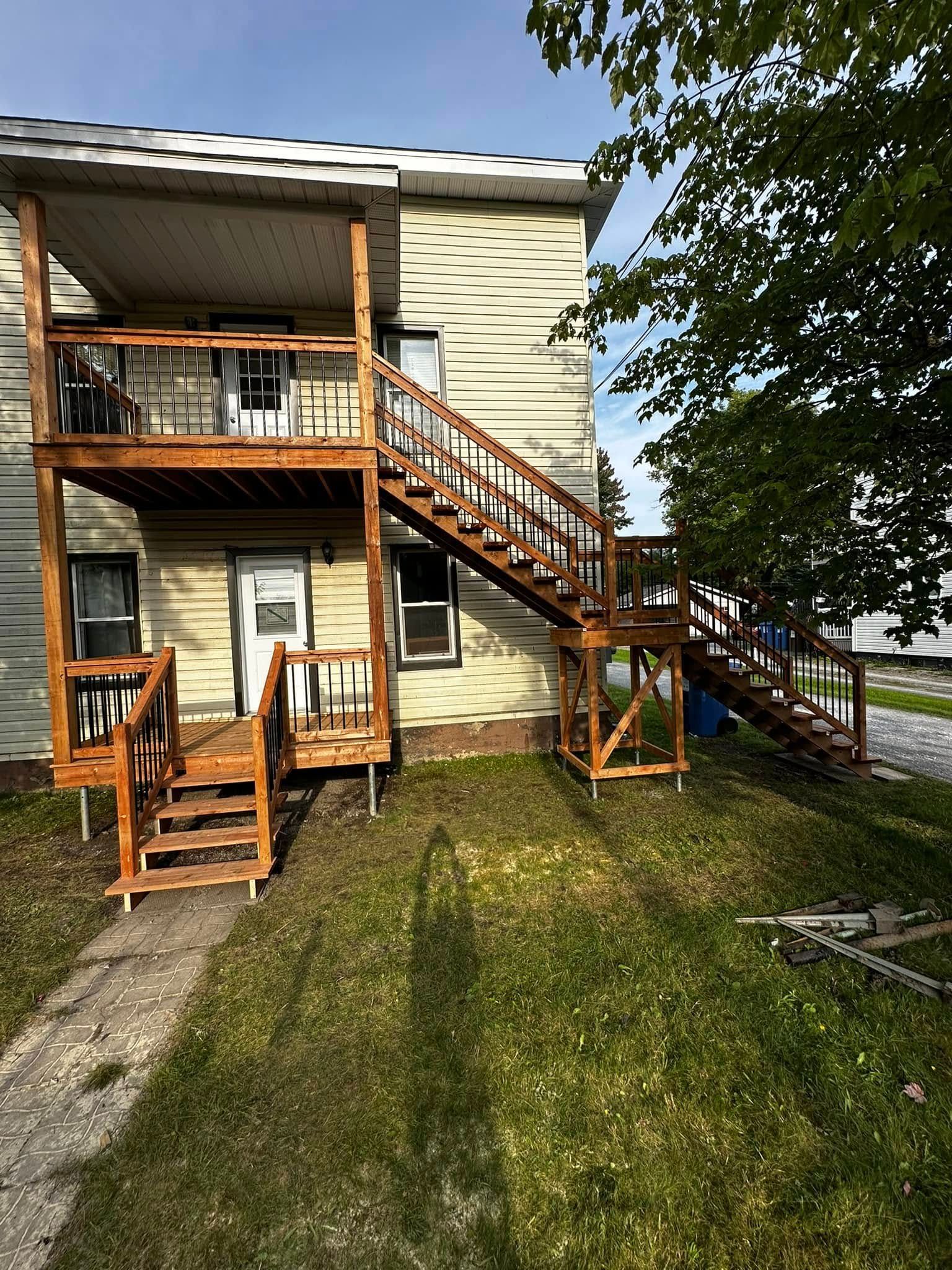 Maison beige à deux étages avec balcons en bois, escalier extérieur et cour gazonnée sous un ciel bleu.