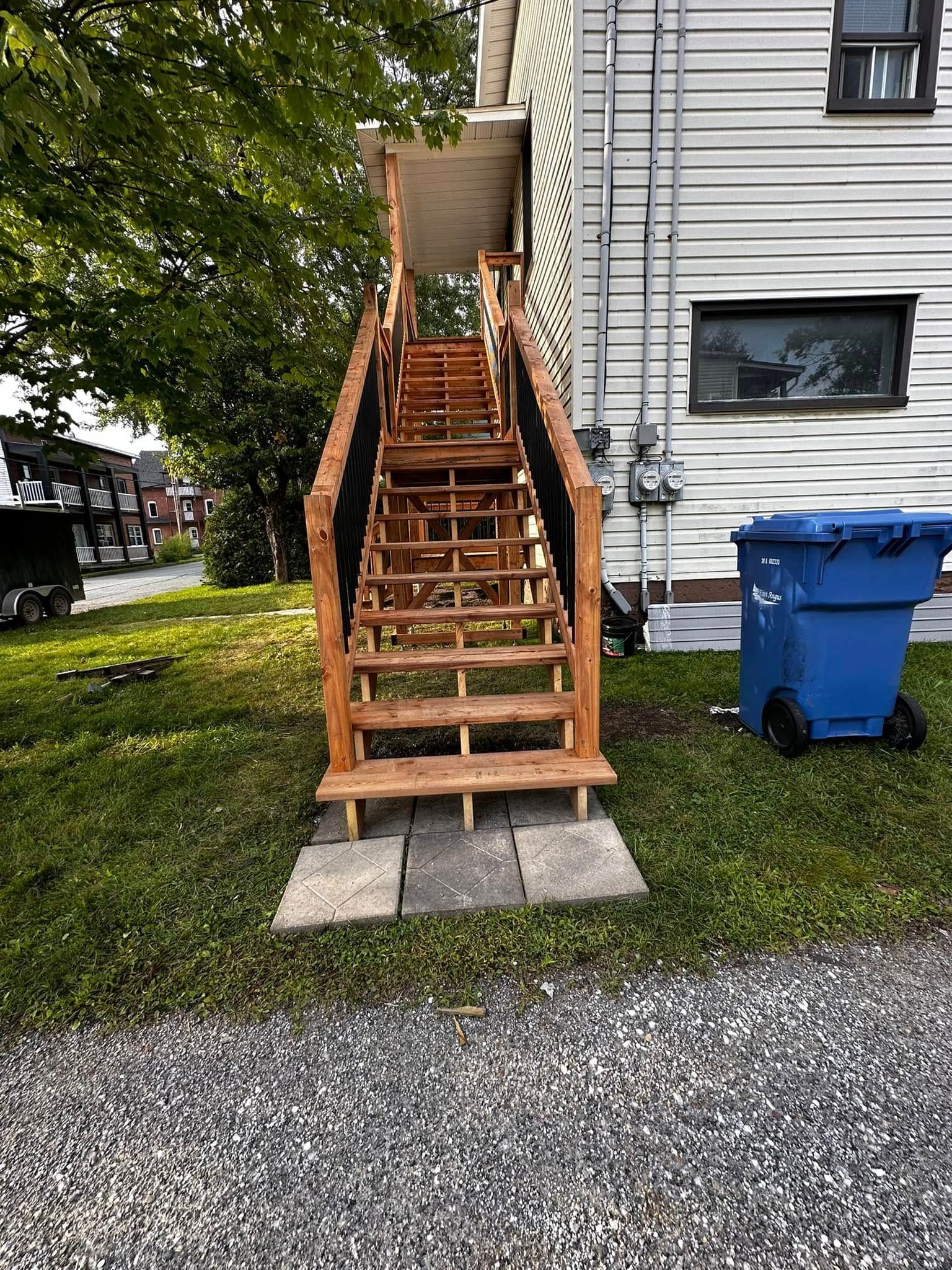 Un escalier extérieur en bois se trouve à côté d'une maison, avec une poubelle de recyclage bleue à droite et de l'herbe en contrebas.