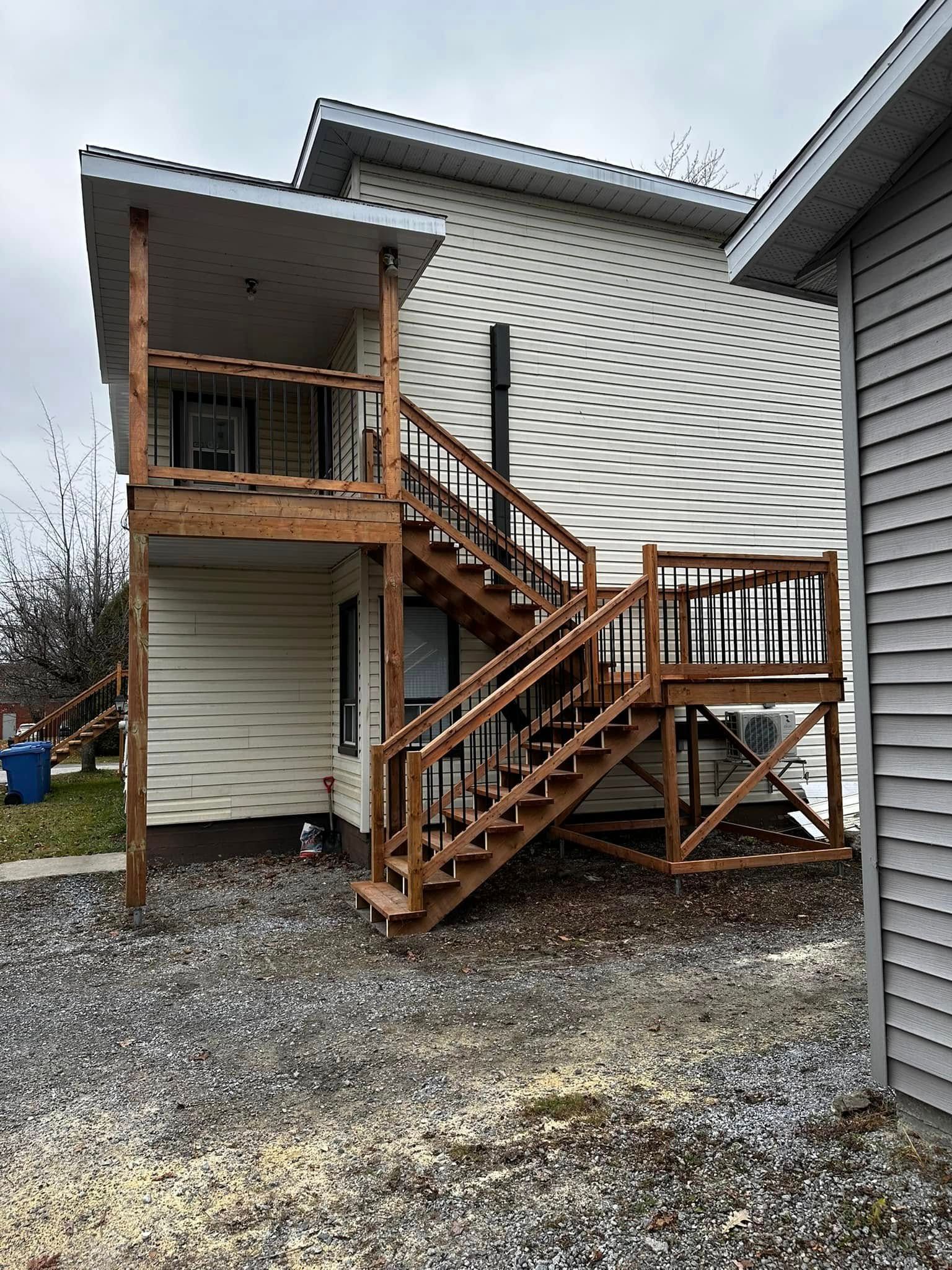 Escalier extérieur en bois et terrasse attenants à une maison blanche sur un terrain gravillonné.