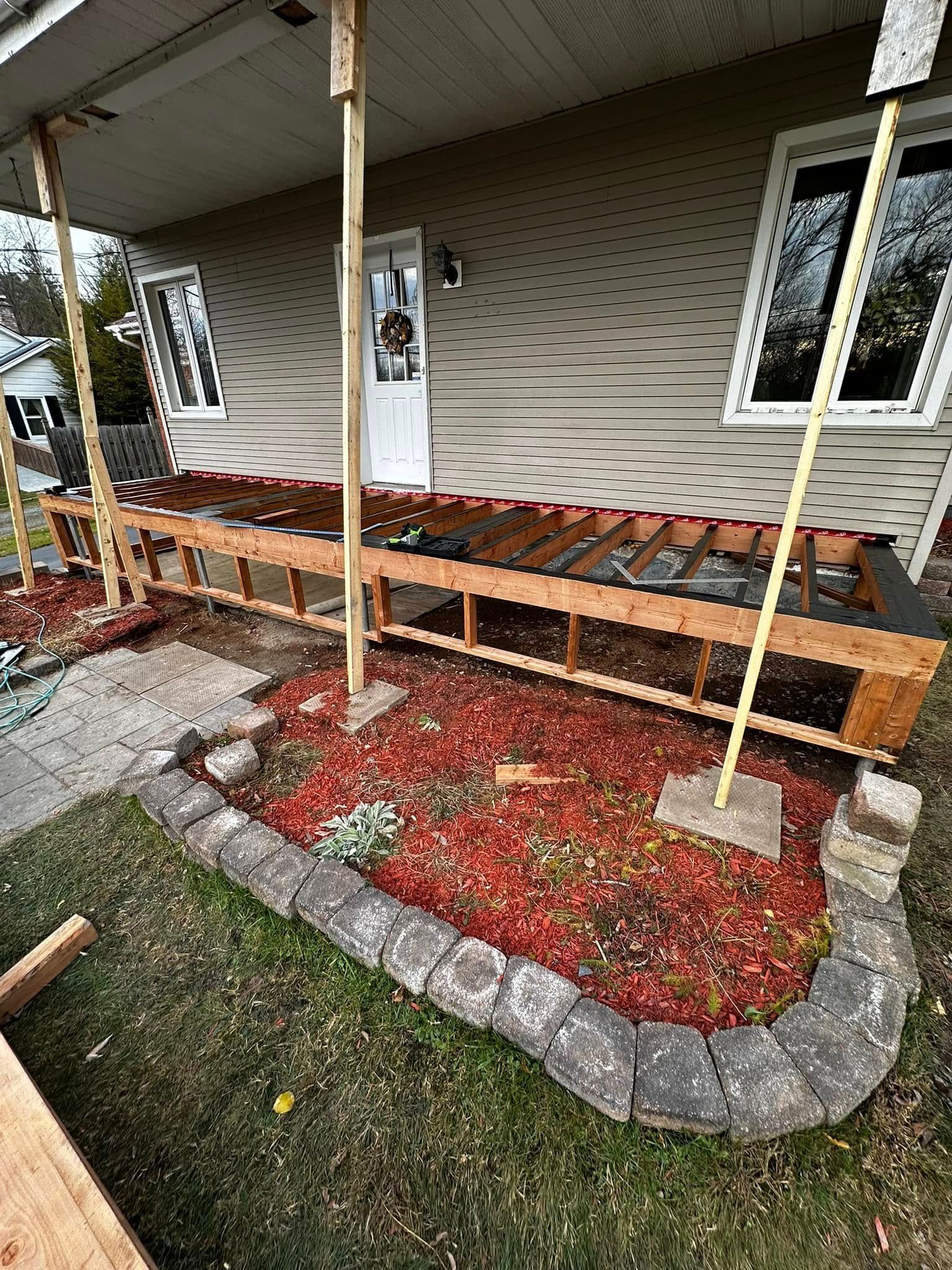 Porche d'entrée endommagé avec marches en bois effondrées, poteaux de soutien et petit parterre aménagé avec paillis rouge.