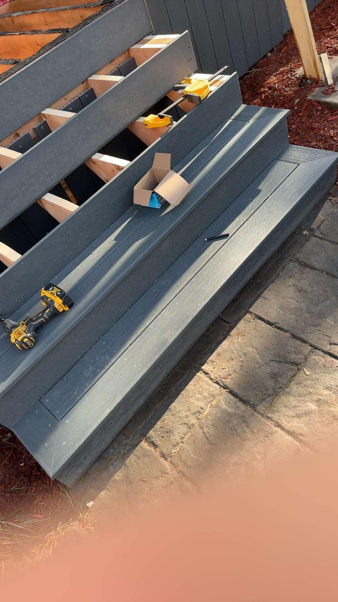 Banc en bois gris avec une petite tasse en carton et un jouet bleu sur les lattes, à l'extérieur sur une terrasse en briques.
