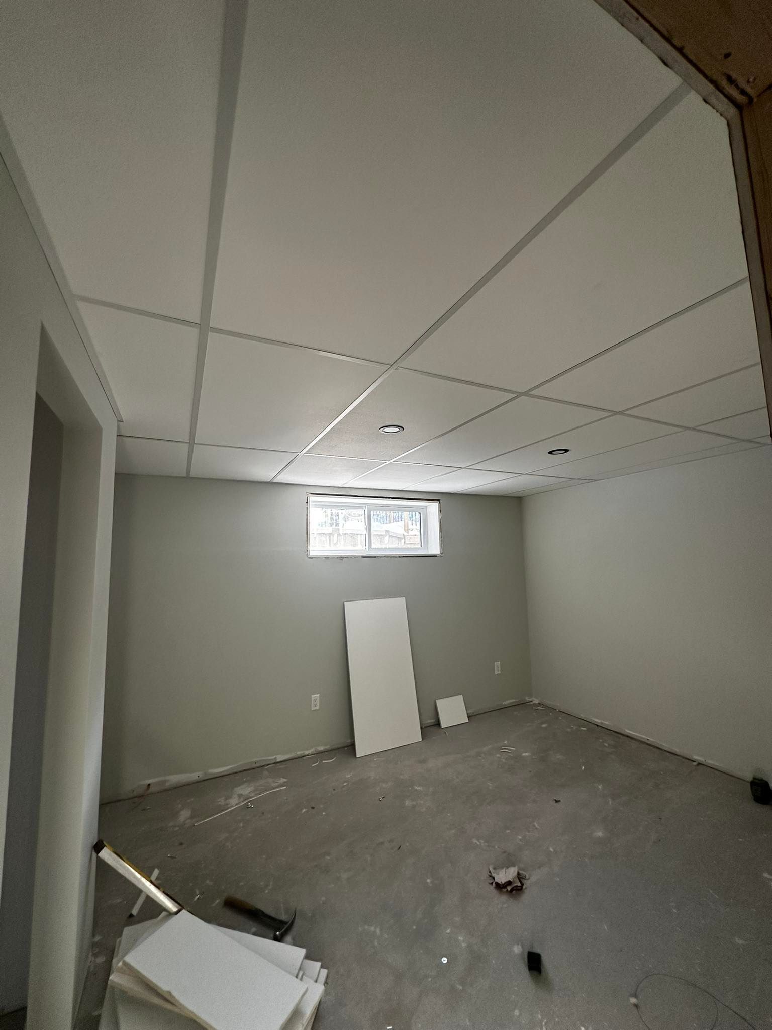 Pièce vide et inachevée, jonchée de débris, aux murs gris, avec un faux plafond et une petite fenêtre.