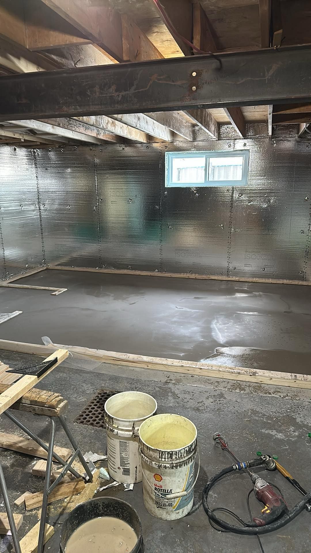 Sous-sol en travaux avec sol humide, poutres apparentes, seaux et une petite fenêtre.