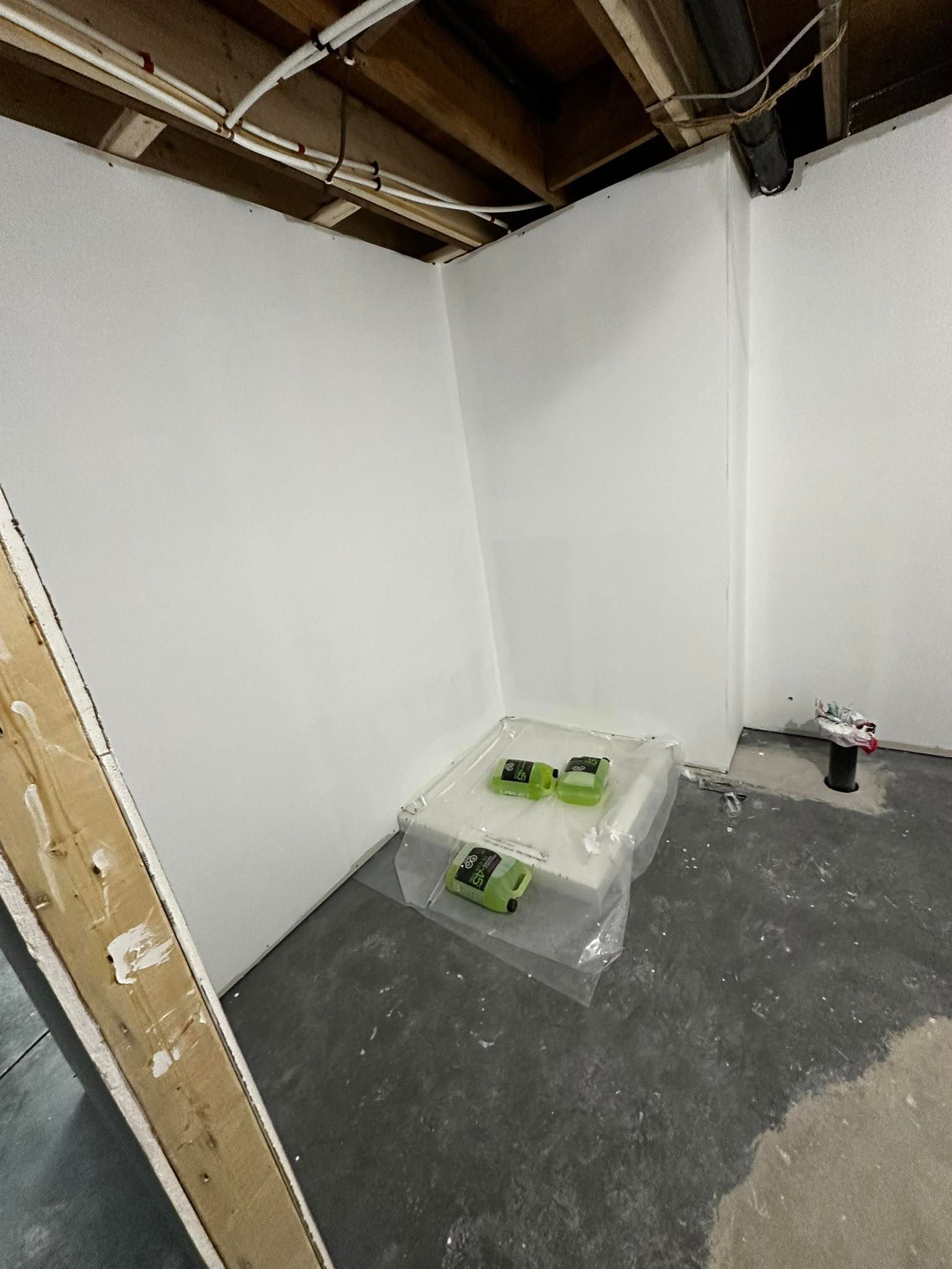 Coin rénovation avec murs blancs, poutres apparentes au plafond et outils au sol près d'un objet recouvert de plastique