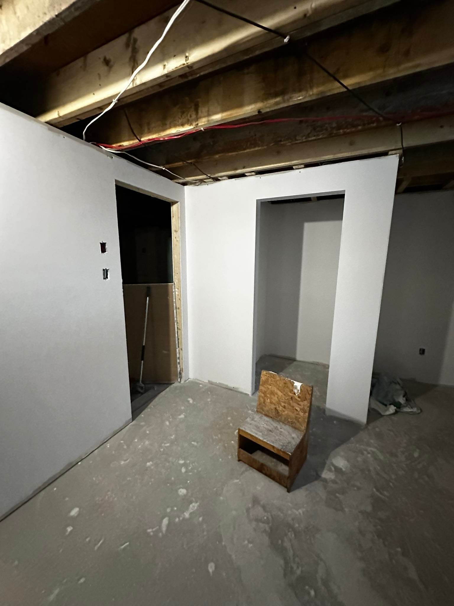Pièce inachevée avec des murs blancs, des poutres apparentes au plafond et une petite chaise en bois sur un sol en béton poussiéreux.