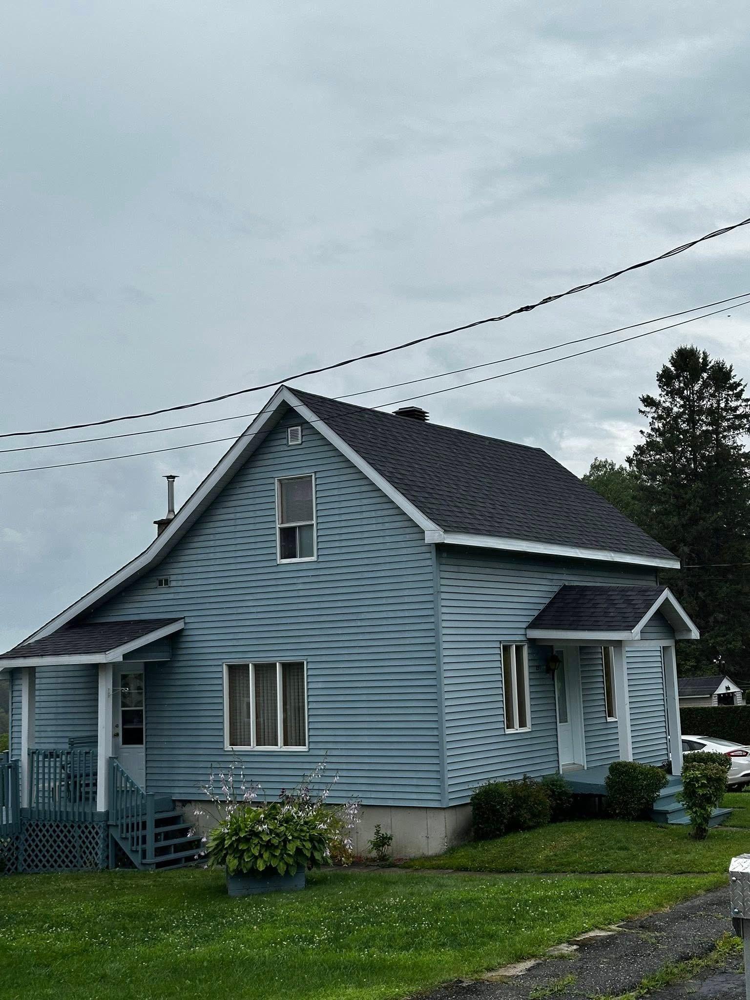 Maison bleue à deux étages avec un toit sombre et une petite véranda sur un terrain herbeux sous un ciel nuageux