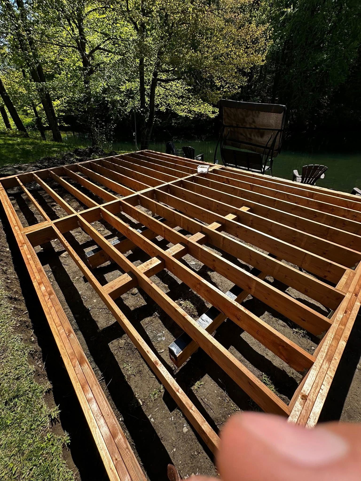 La structure d'une terrasse en bois est en construction à l'extérieur, avec les solives disposées dans un jardin ensoleillé.