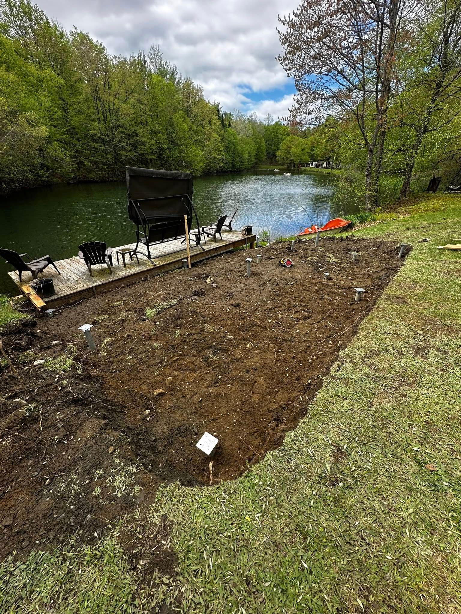 Un parterre de jardin fraîchement débroussaillé au bord d'un étang, avec des arbres verts et une brouette en arrière-plan.