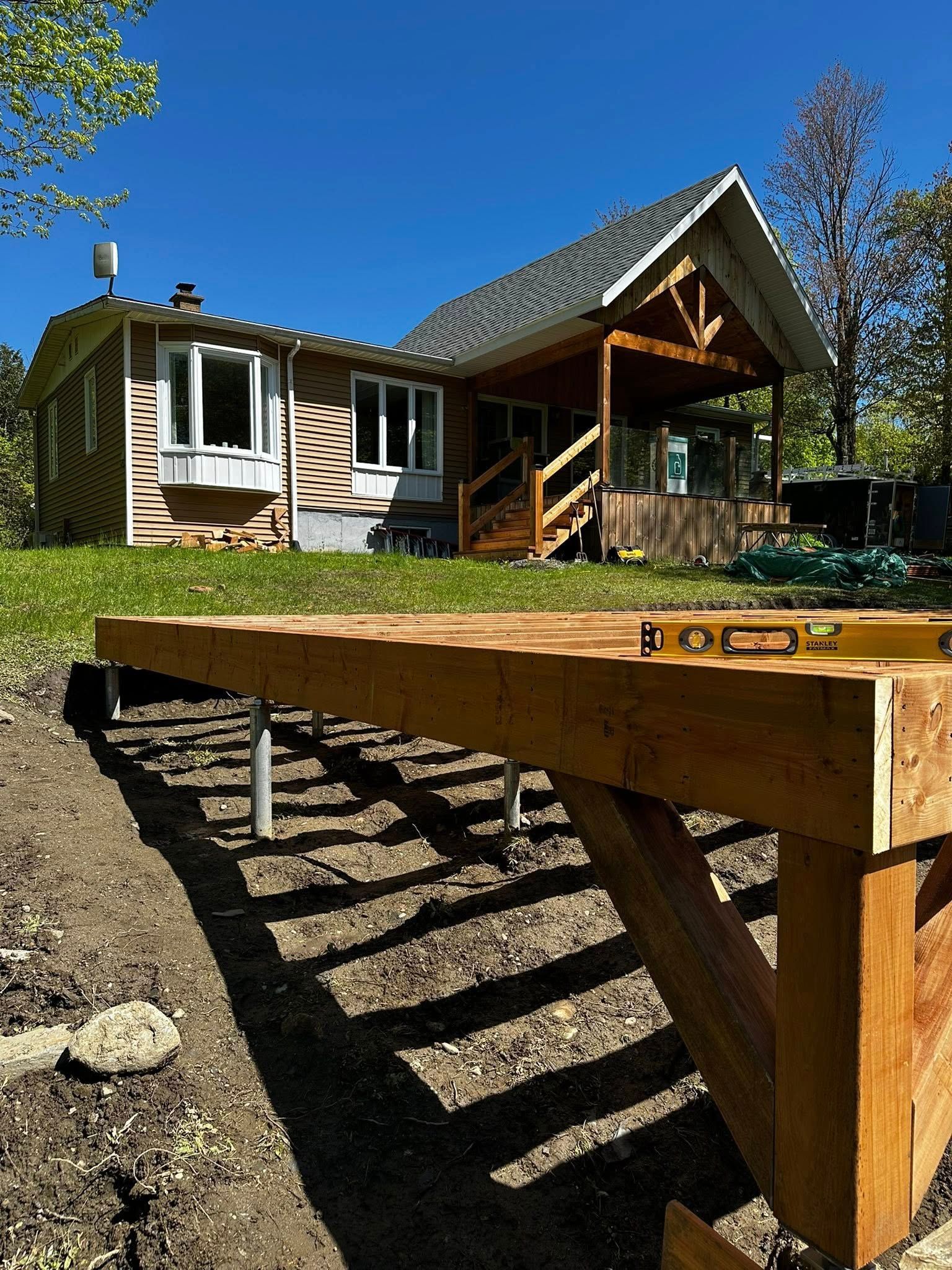 Au premier plan, une structure de terrasse en bois surélevée, avec une maison en construction à l'arrière-plan, par une journée ensoleillée.