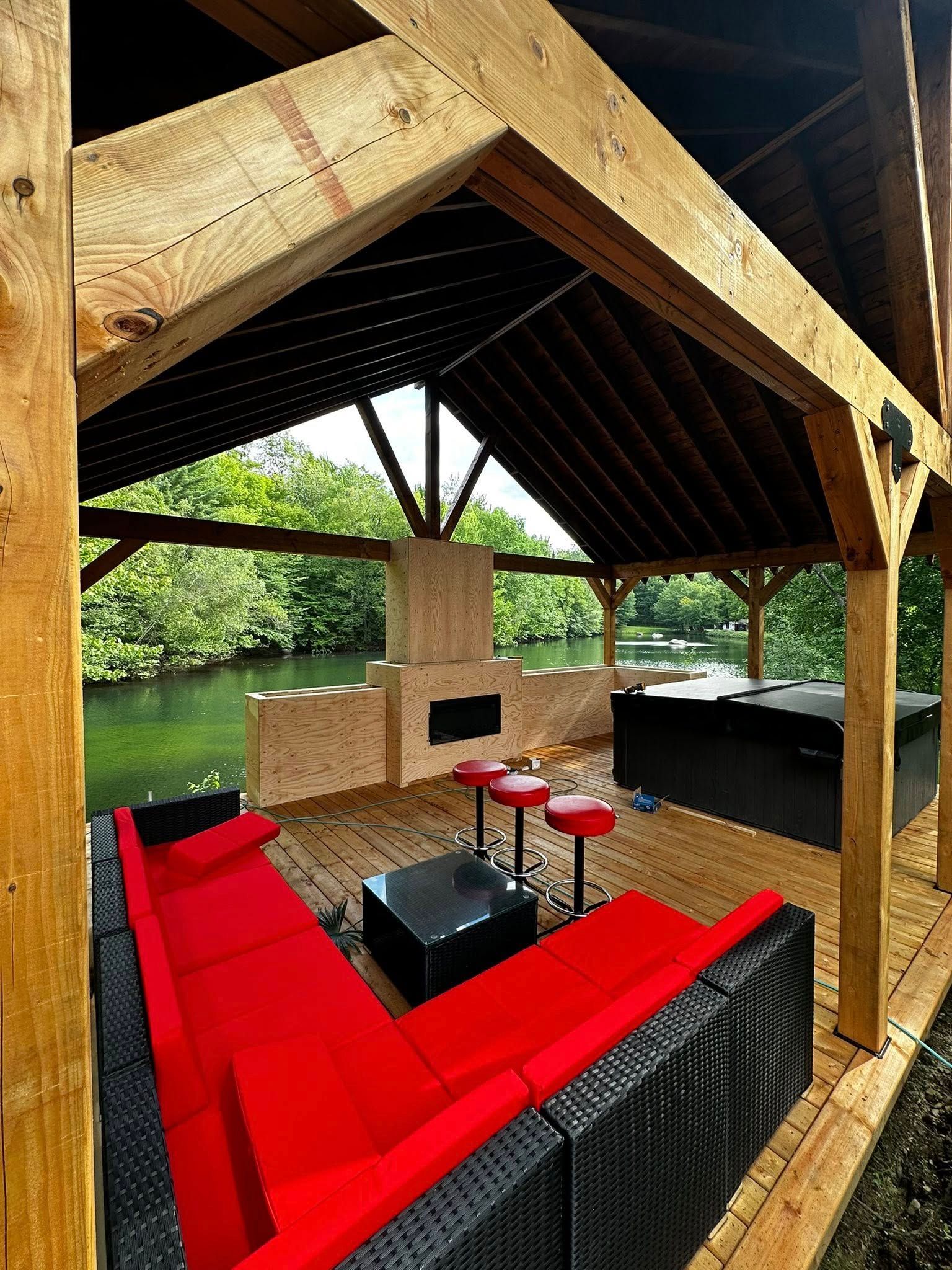 Salon extérieur avec canapé d'angle rouge, cheminée en pierre et vue sur l'eau sous un kiosque en bois