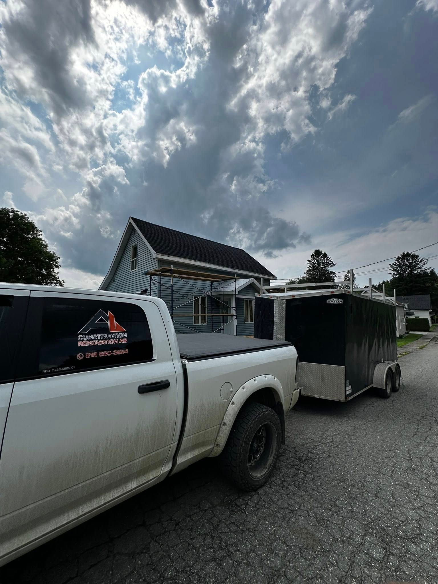 Un pick-up blanc avec une remorque noire garé devant une maison sous un ciel nuageux