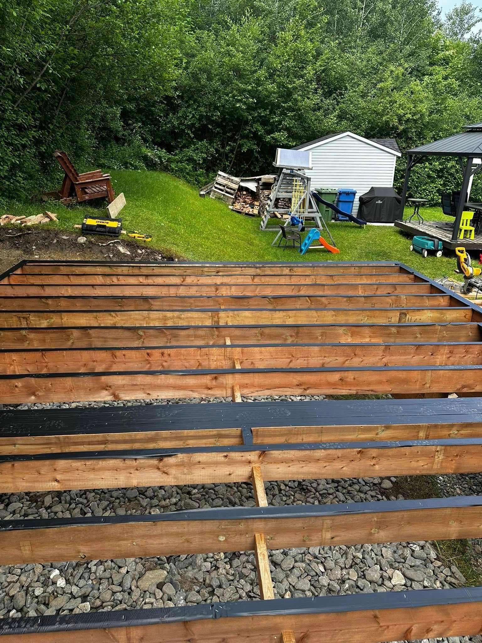 Construction d'une structure de terrasse en bois dans un jardin, avec du gravier, des outils et un abri de jardin en arrière-plan.
