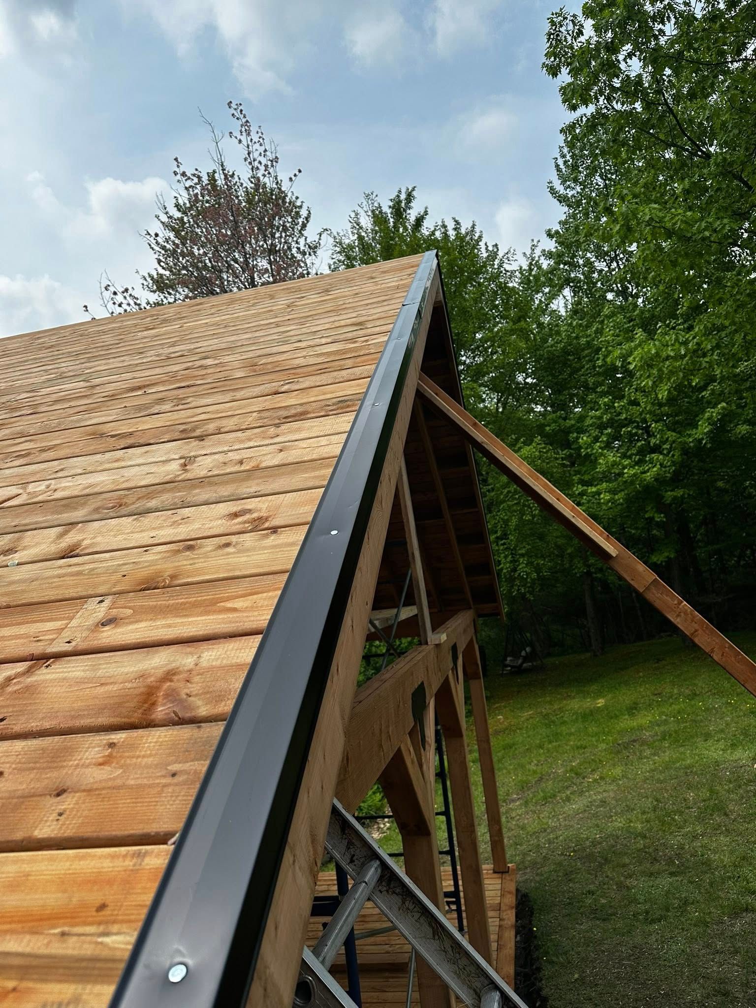 Gros plan sur un toit en bois en construction, avec des finitions noires, se détachant sur des arbres verts et un ciel nuageux.