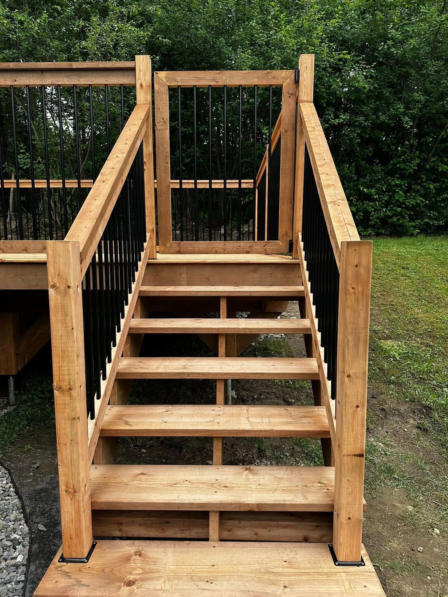 Un escalier extérieur en bois avec rambarde mène à une terrasse, adossée à des haies vertes.