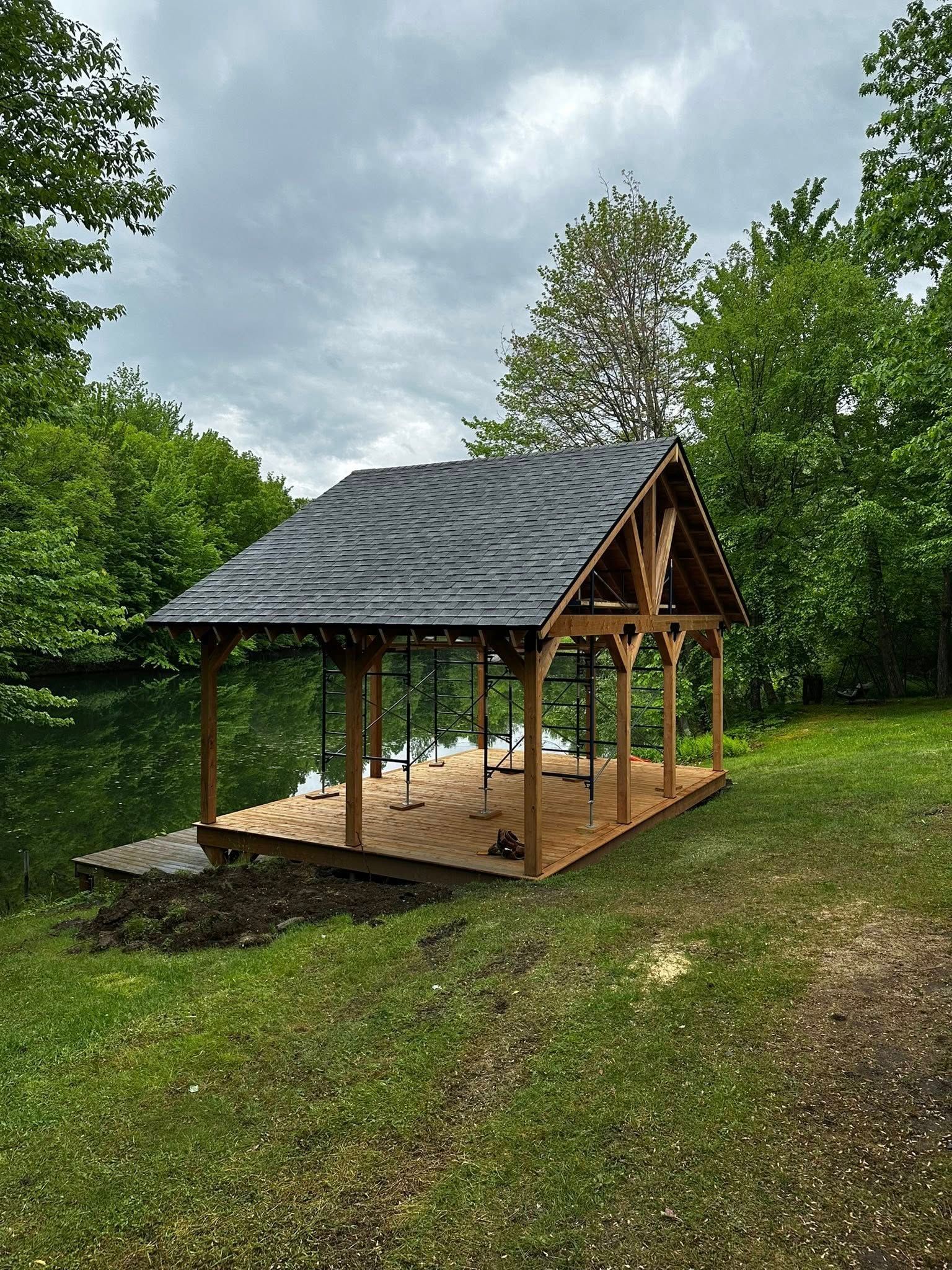 Petit pavillon en bois au toit de bardeaux sombres, dans une cour herbeuse, entouré d'arbres sous un ciel nuageux.