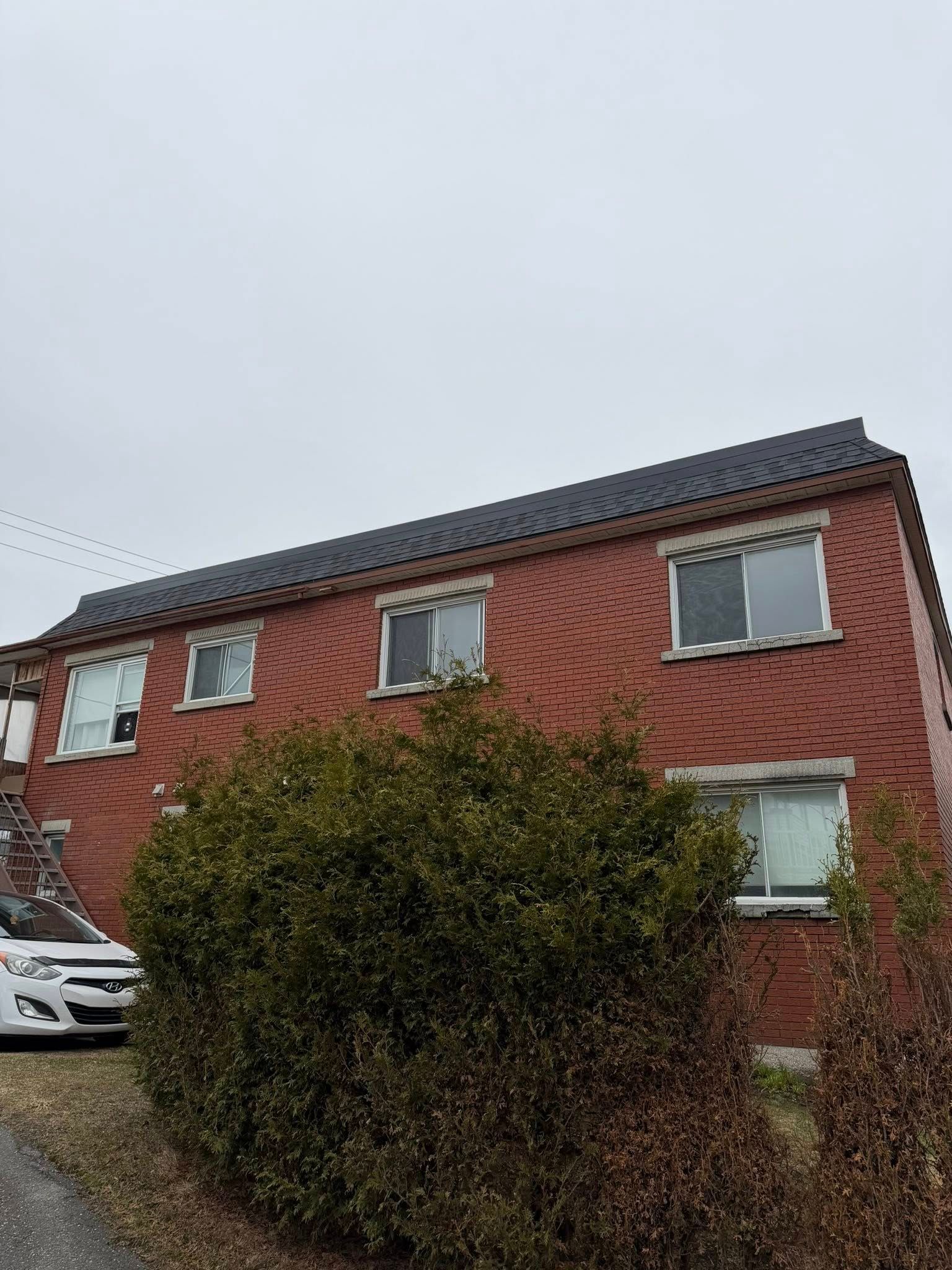 Maison de deux étages en briques rouges au toit sombre, sous un ciel couvert, partiellement dissimulée par un grand buisson.