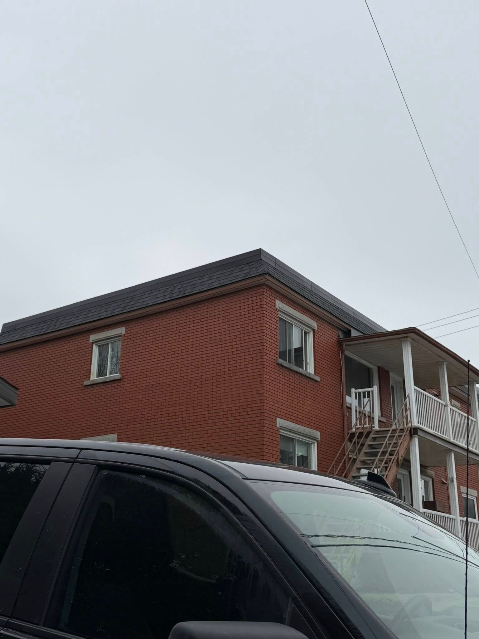Bâtiment en briques rouges aux balcons blancs, derrière une voiture noire par temps couvert.