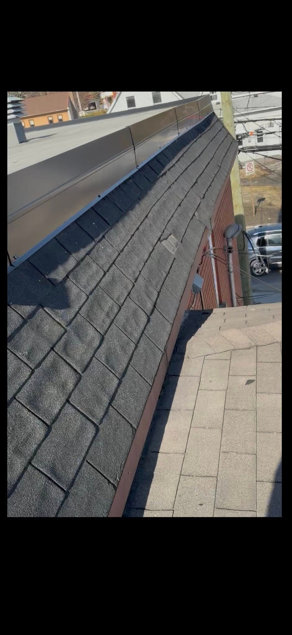 Vue depuis le toit d'une toiture en bardeaux noirs, jouxtant un mur beige et des bâtiments de la ville en contrebas.
