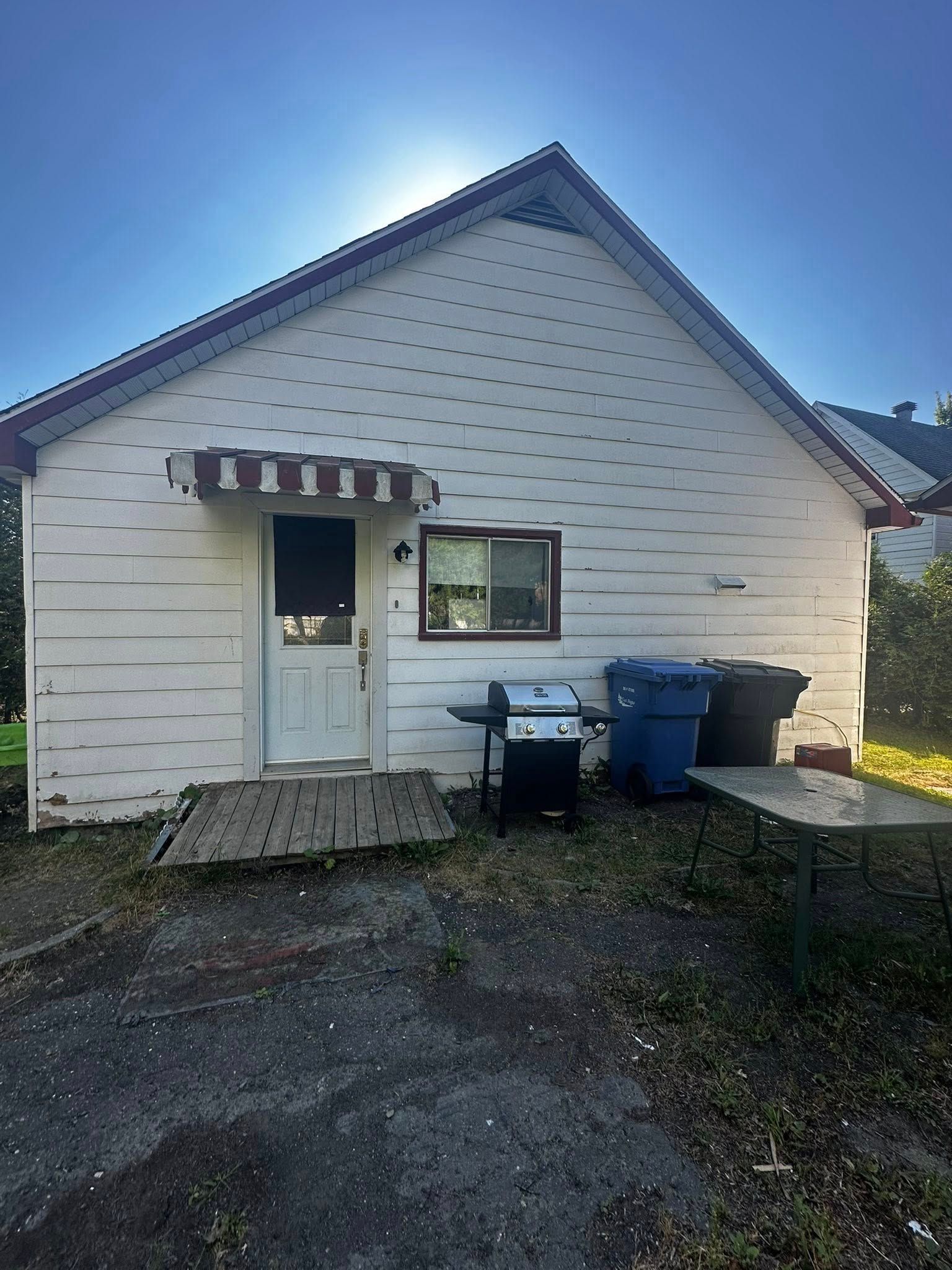 Extérieur d'une petite maison blanche avec auvent, porte d'entrée, fenêtre et poubelles, au bord d'une allée de gravier.