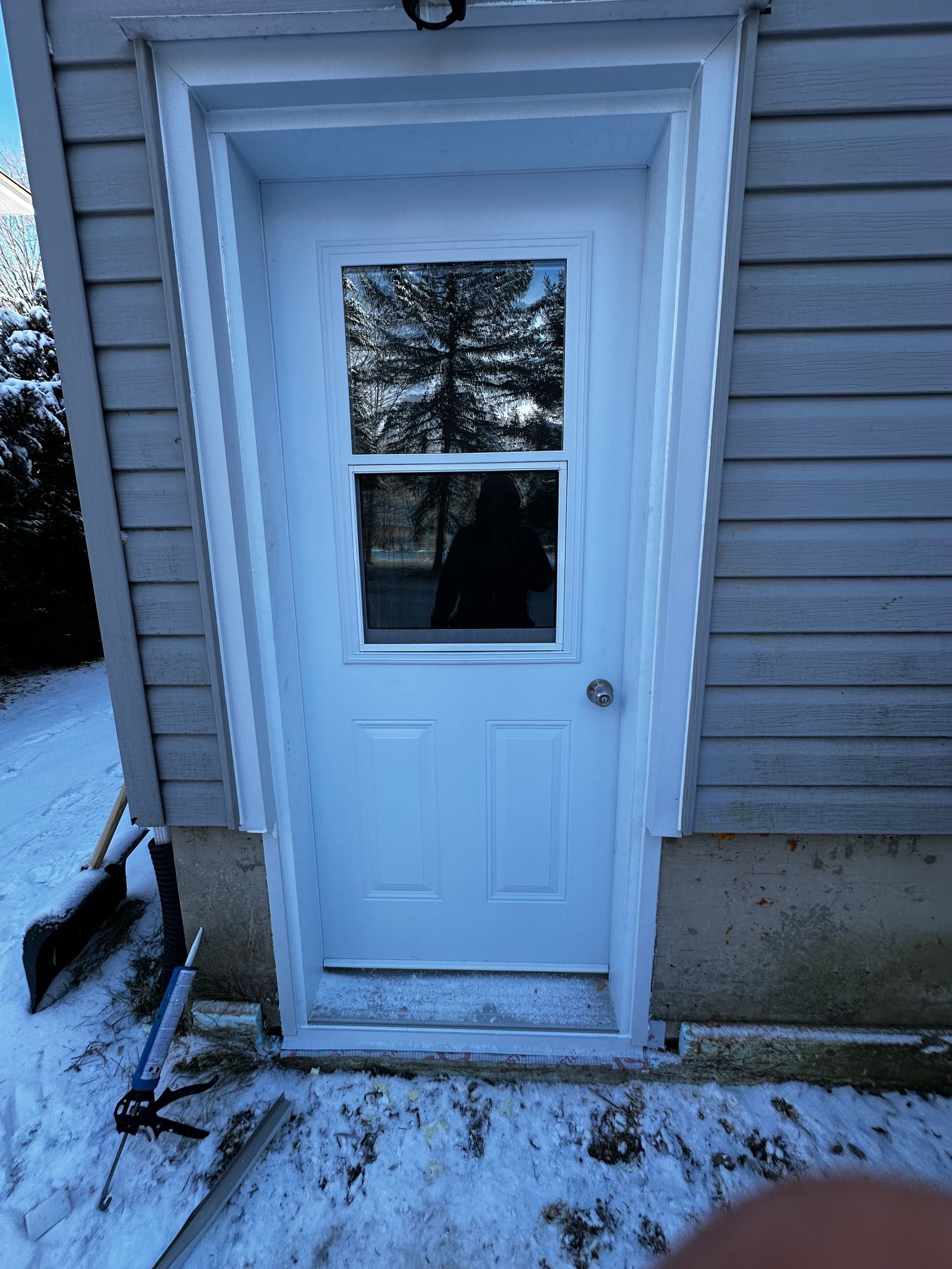 Porte extérieure blanche avec une fenêtre, encadrée par un bardage beige et de la neige au sol