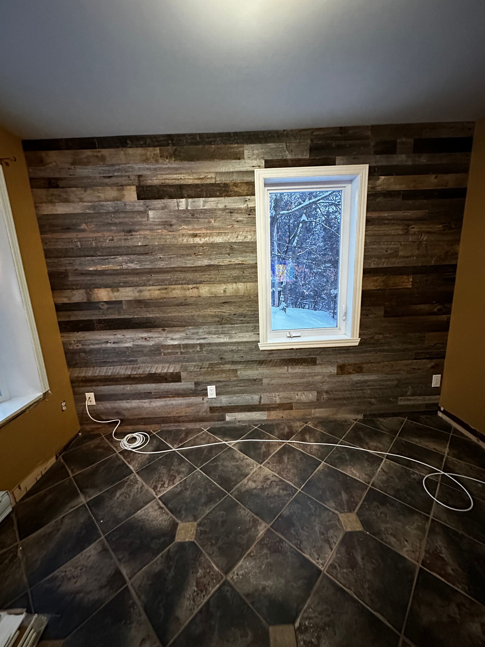 Pièce vide avec un mur d'accent en bois rustique, une fenêtre centrale et un sol carrelé foncé.