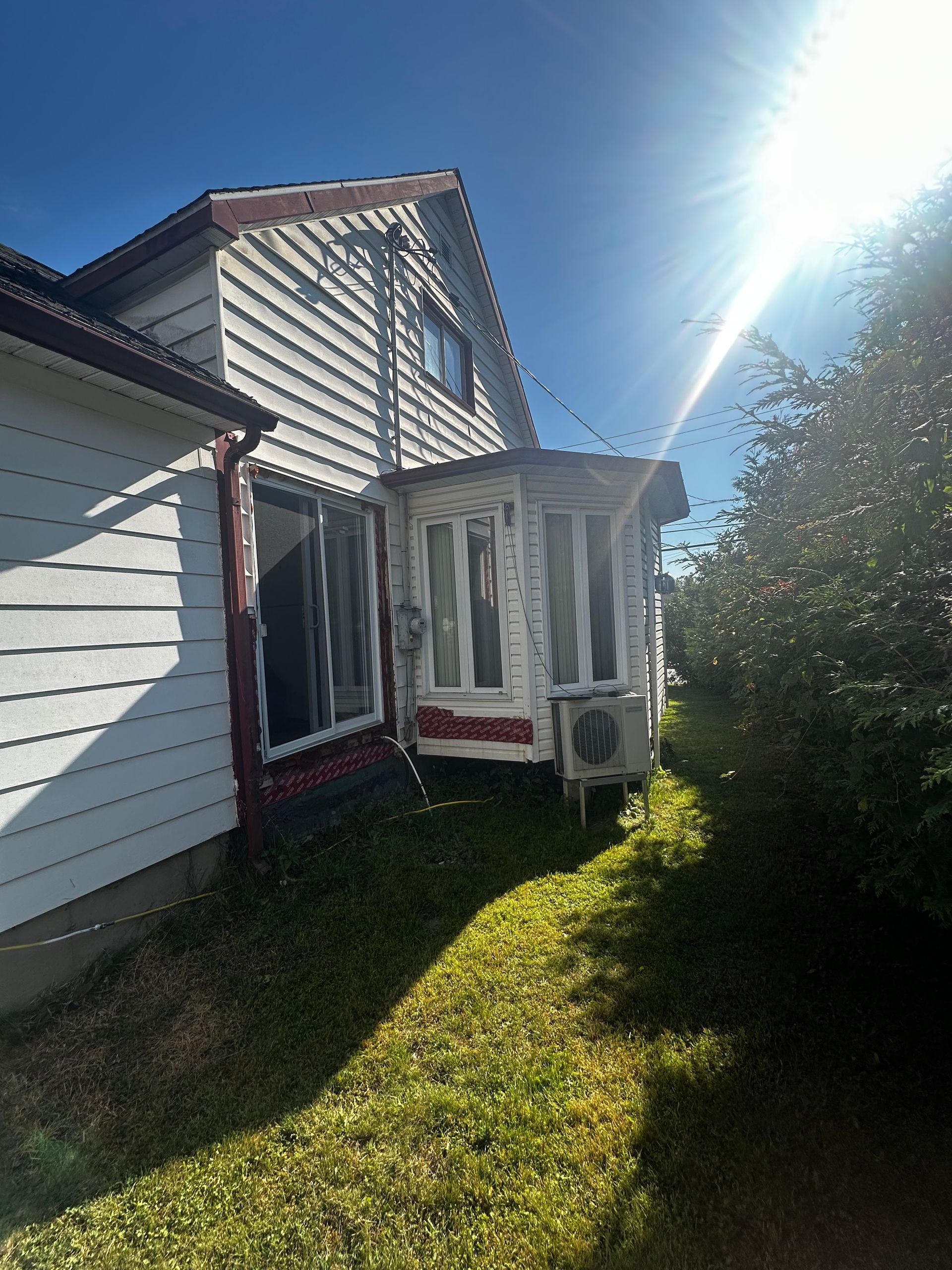 Façade blanche d'une maison avec fenêtres à encadrement rouge, porche et climatiseur, baignée de soleil.