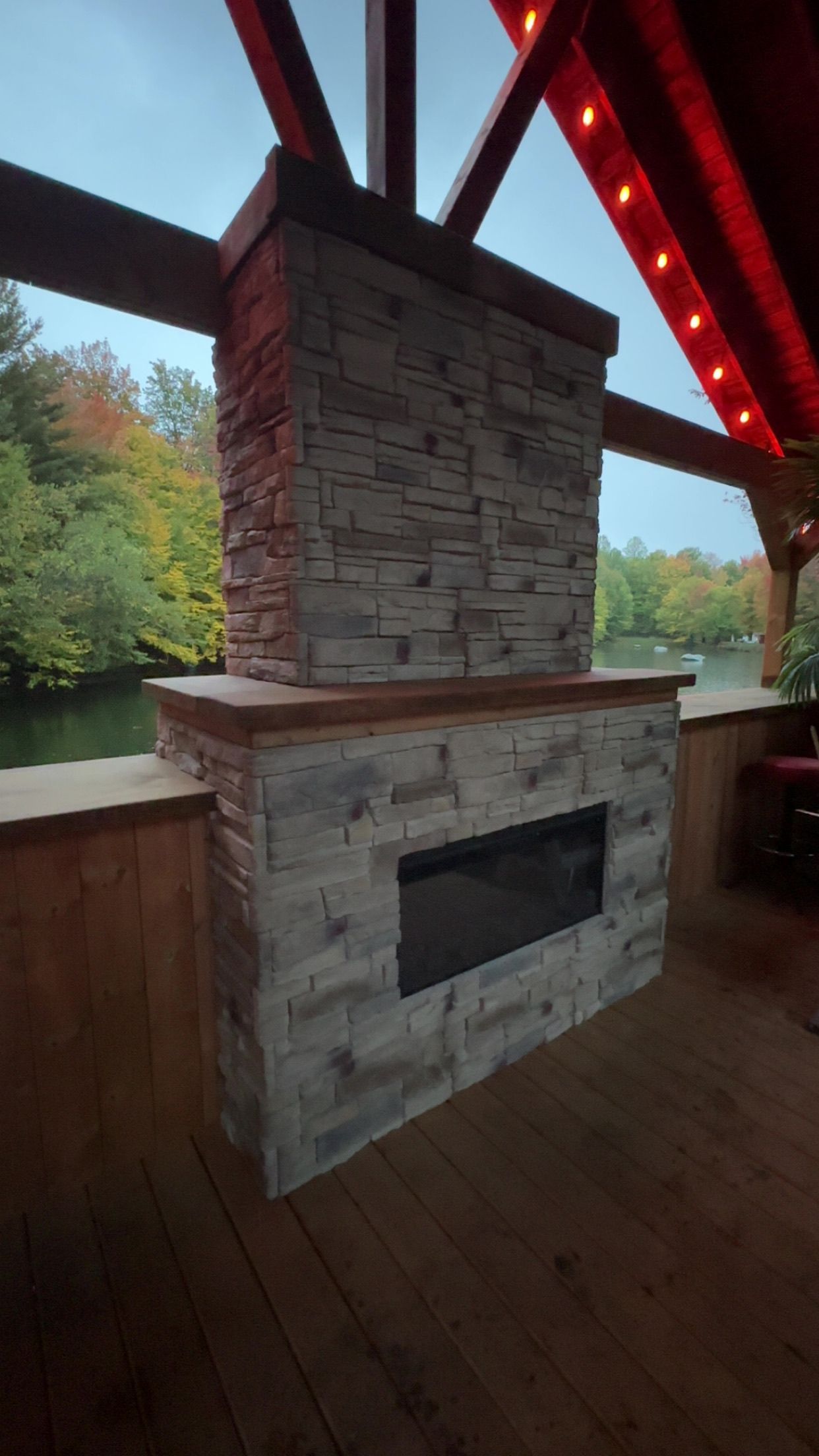 Cheminée en pierre avec insert noir sur une terrasse en bois sous un pavillon au toit rouge, avec des arbres en arrière-plan