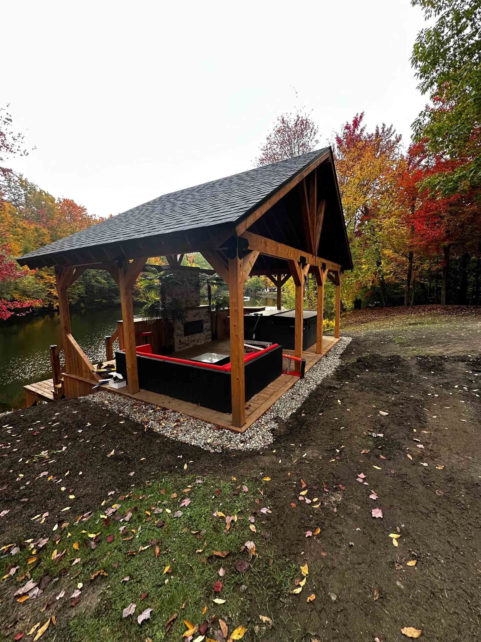 Un kiosque en bois au toit de bardeaux sombres, sur un terrain en terre battue, entouré d'arbres d'automne et de feuilles mortes.