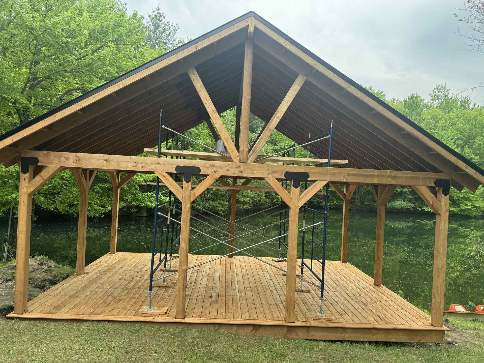 Pavillon en bois à ciel ouvert avec un toit pointu sur un site boisé, sol non fini et poteaux de soutien.