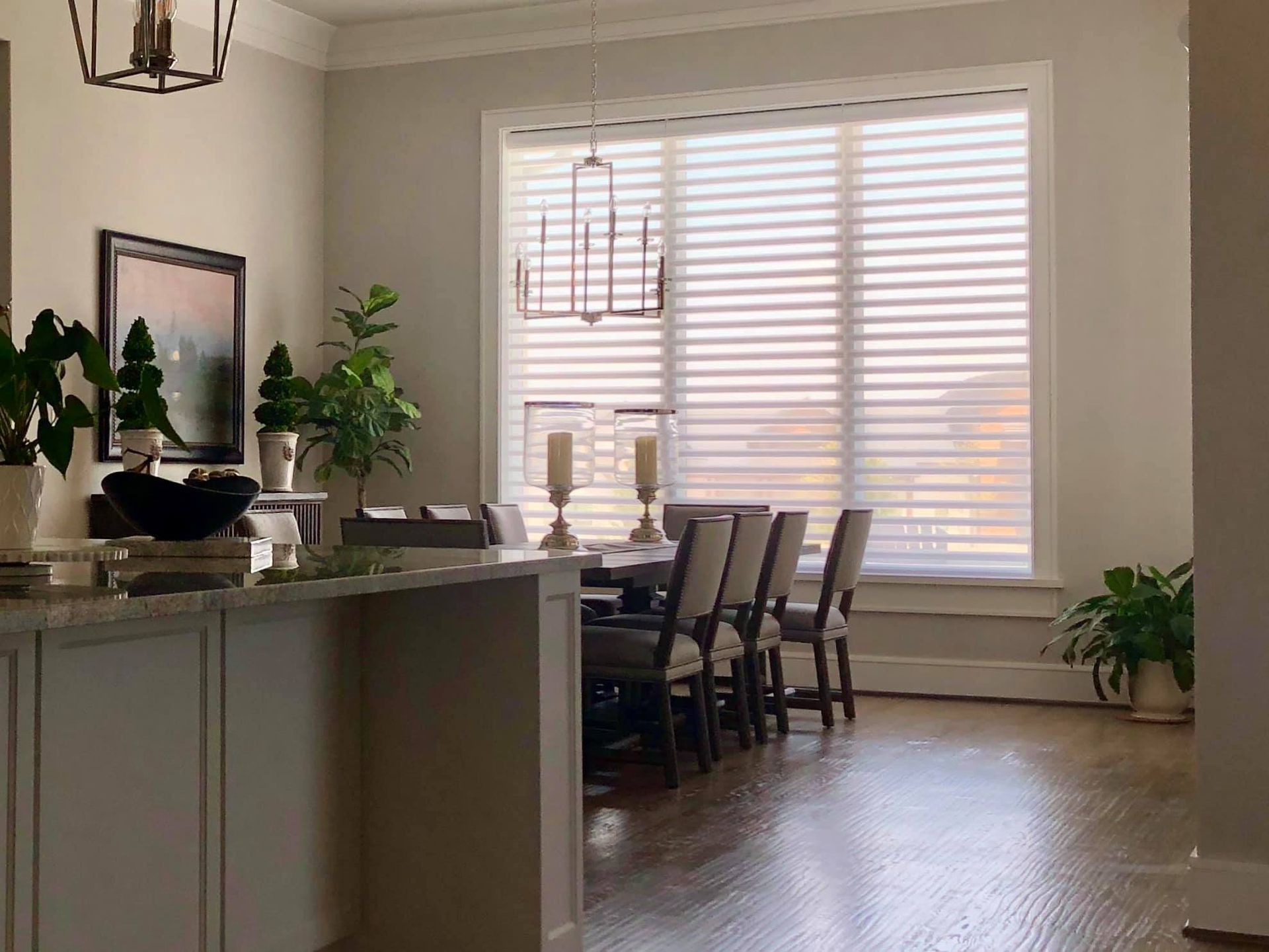 Dining room with a table set for a meal, large window with blinds, plants, and a chandelier.