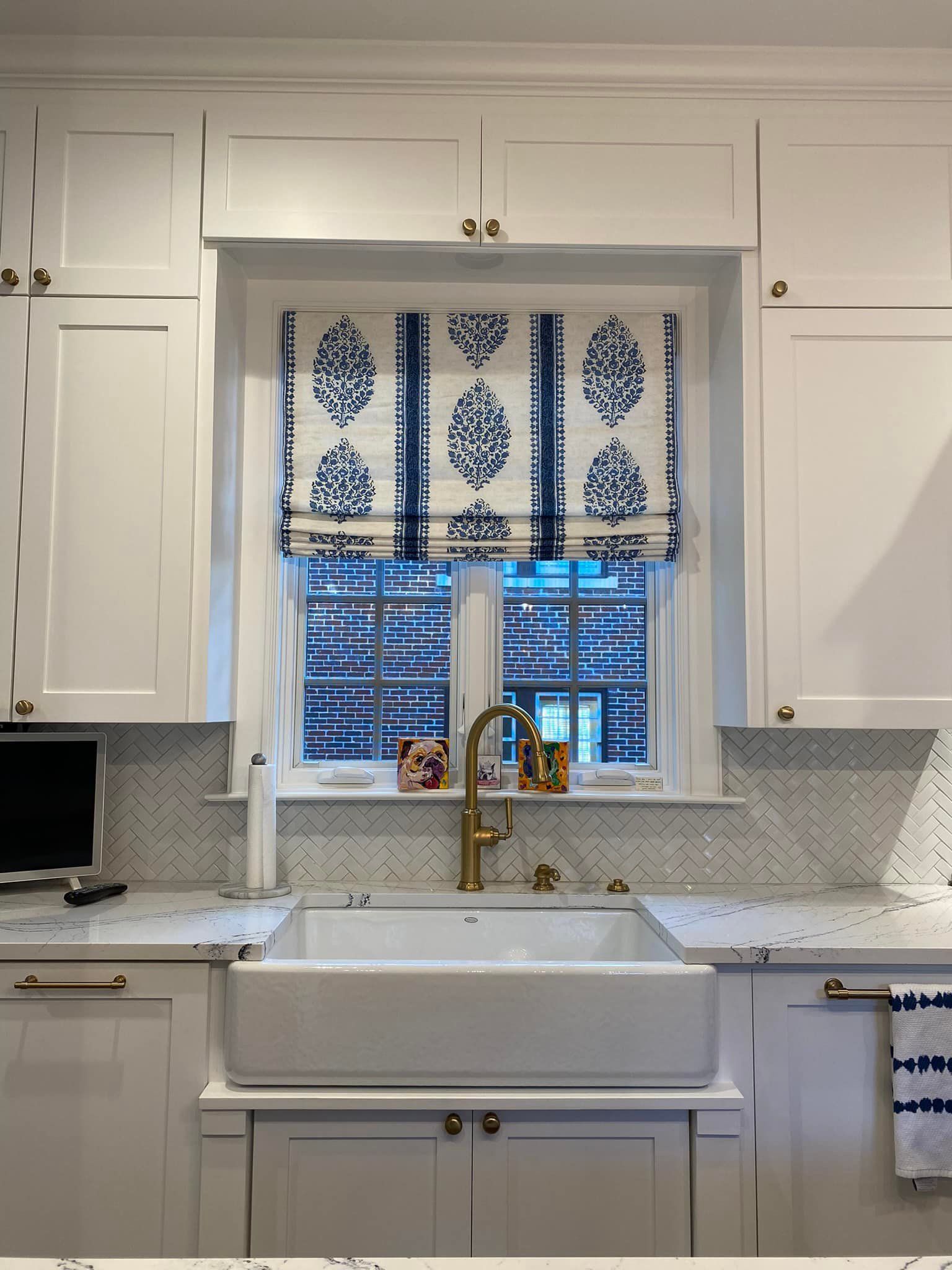 Kitchen with white cabinets, farmhouse sink, blue and white patterned window shade, gold faucet.