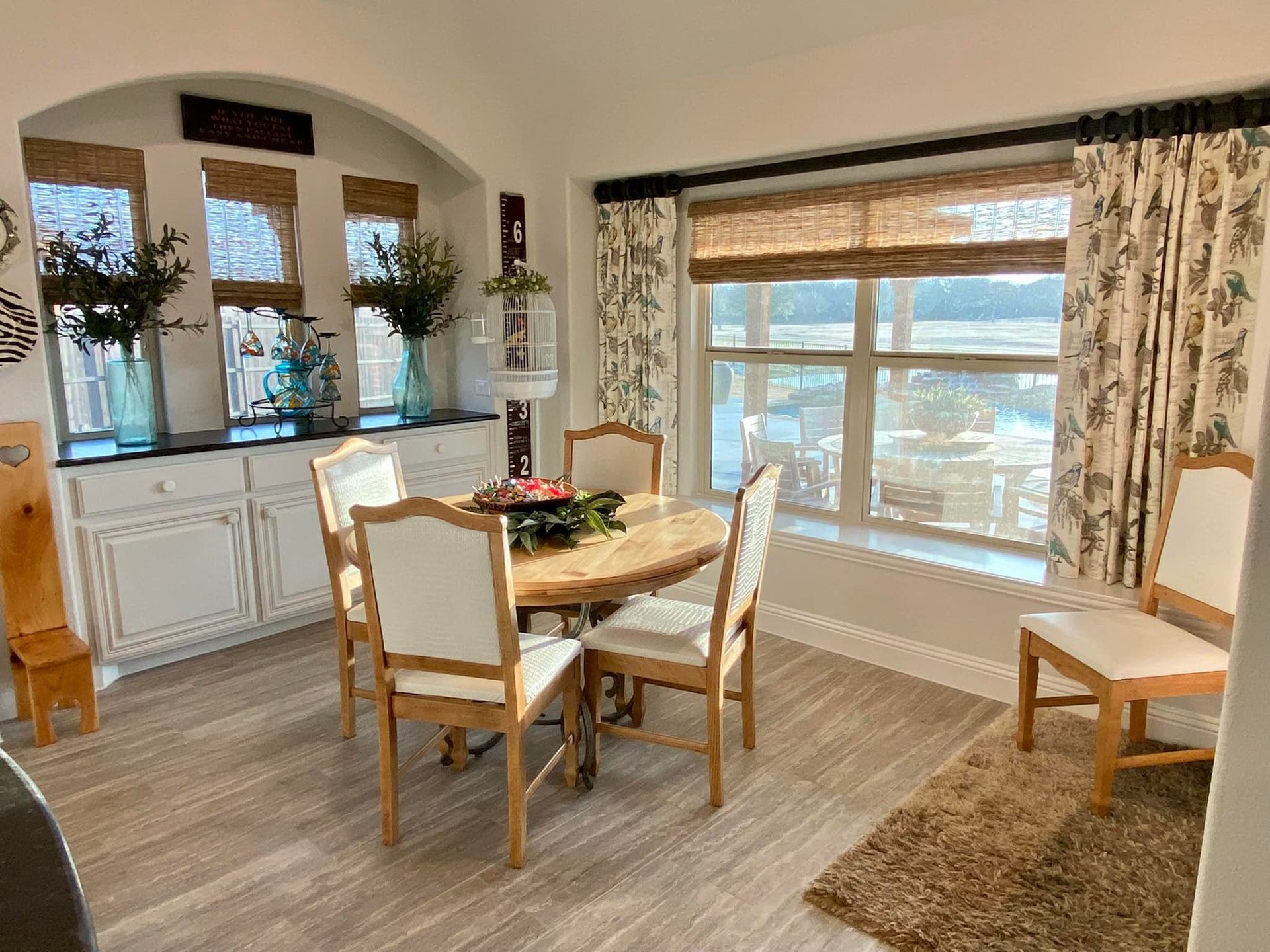 Dining room with oval table, chairs, and window with shades and floral curtains.