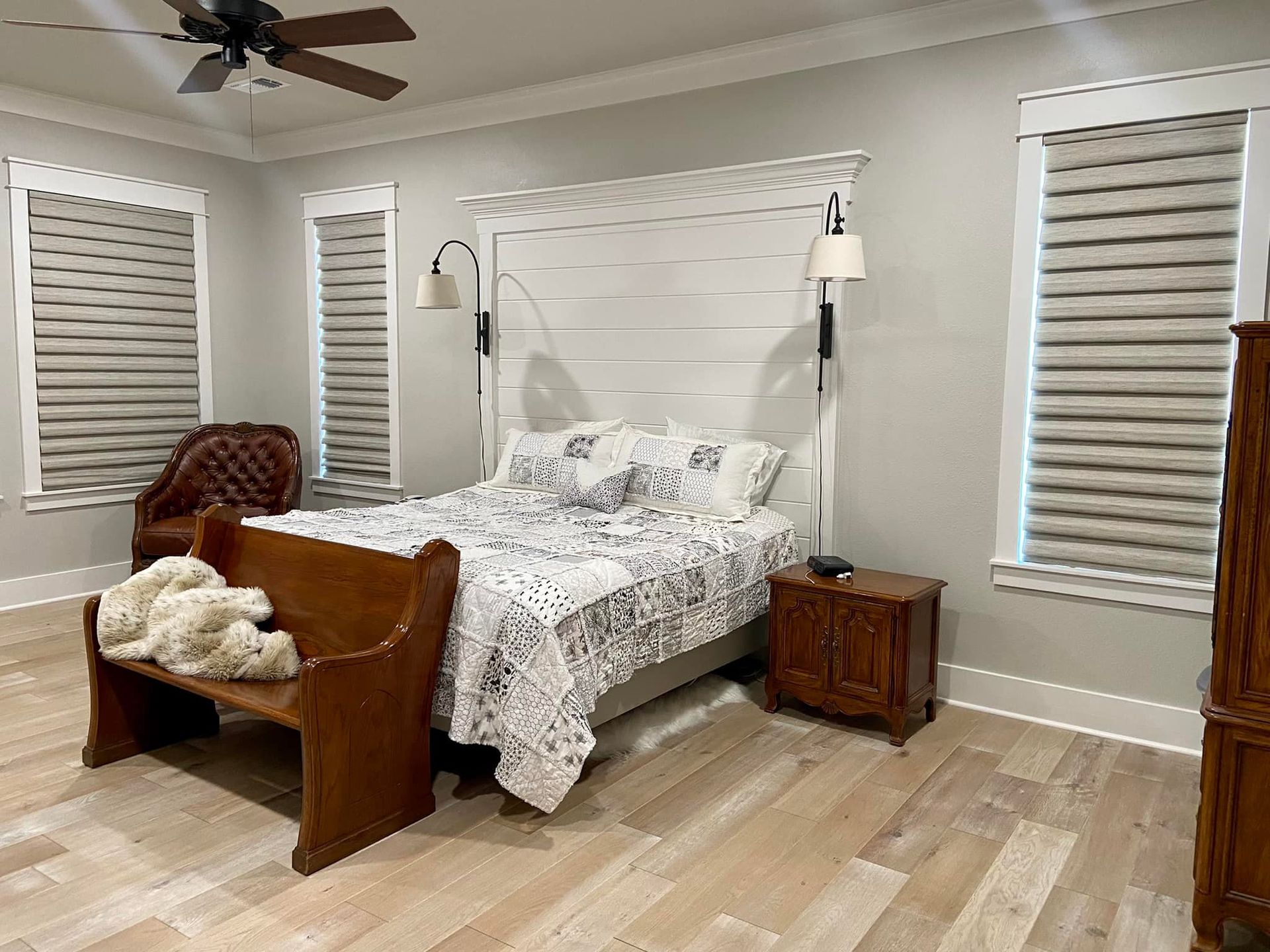 Bedroom with light wood floor, bed with white headboard, bench, shades on windows.