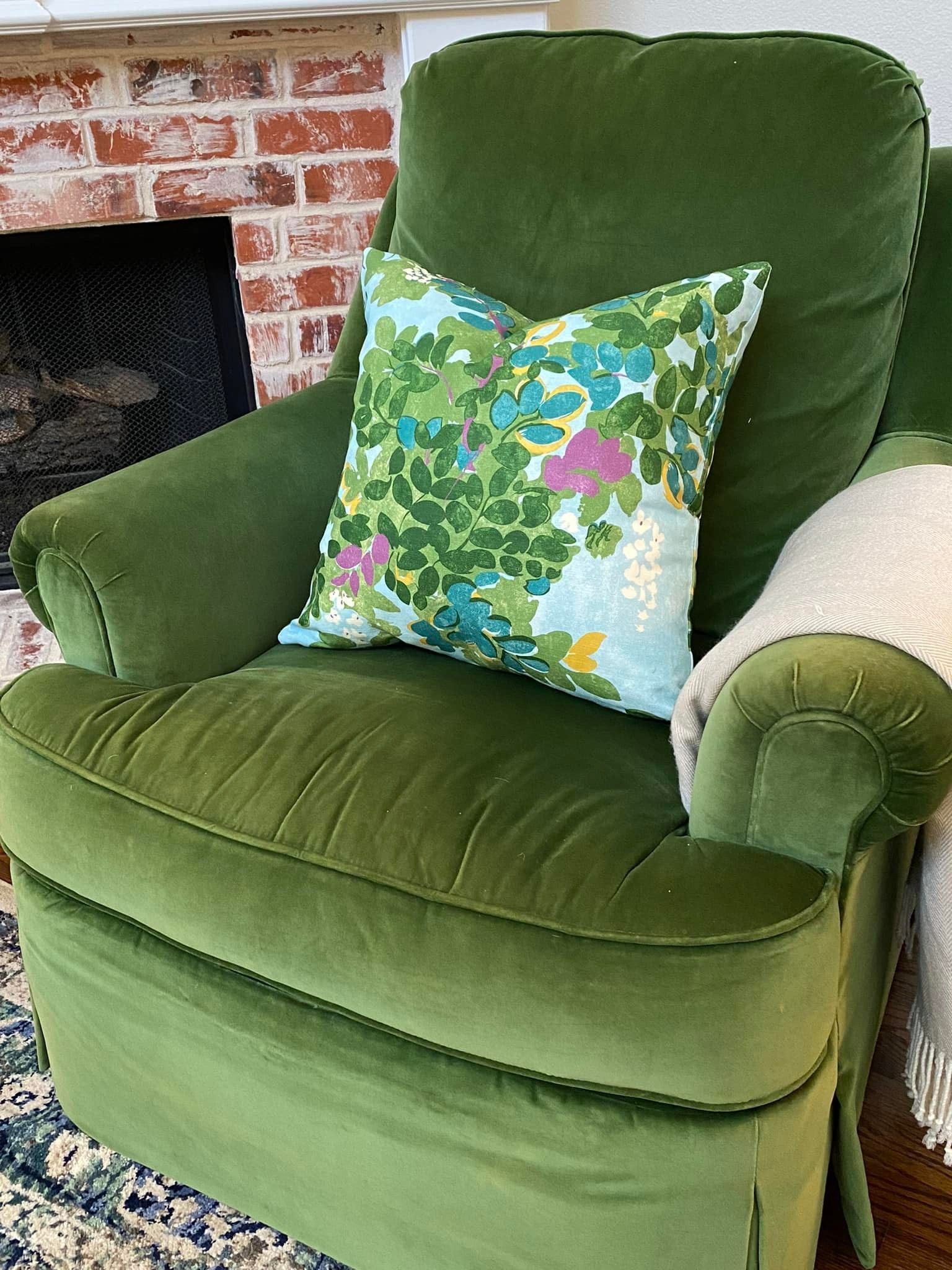 Green velvet armchair with floral pillow near a brick fireplace.