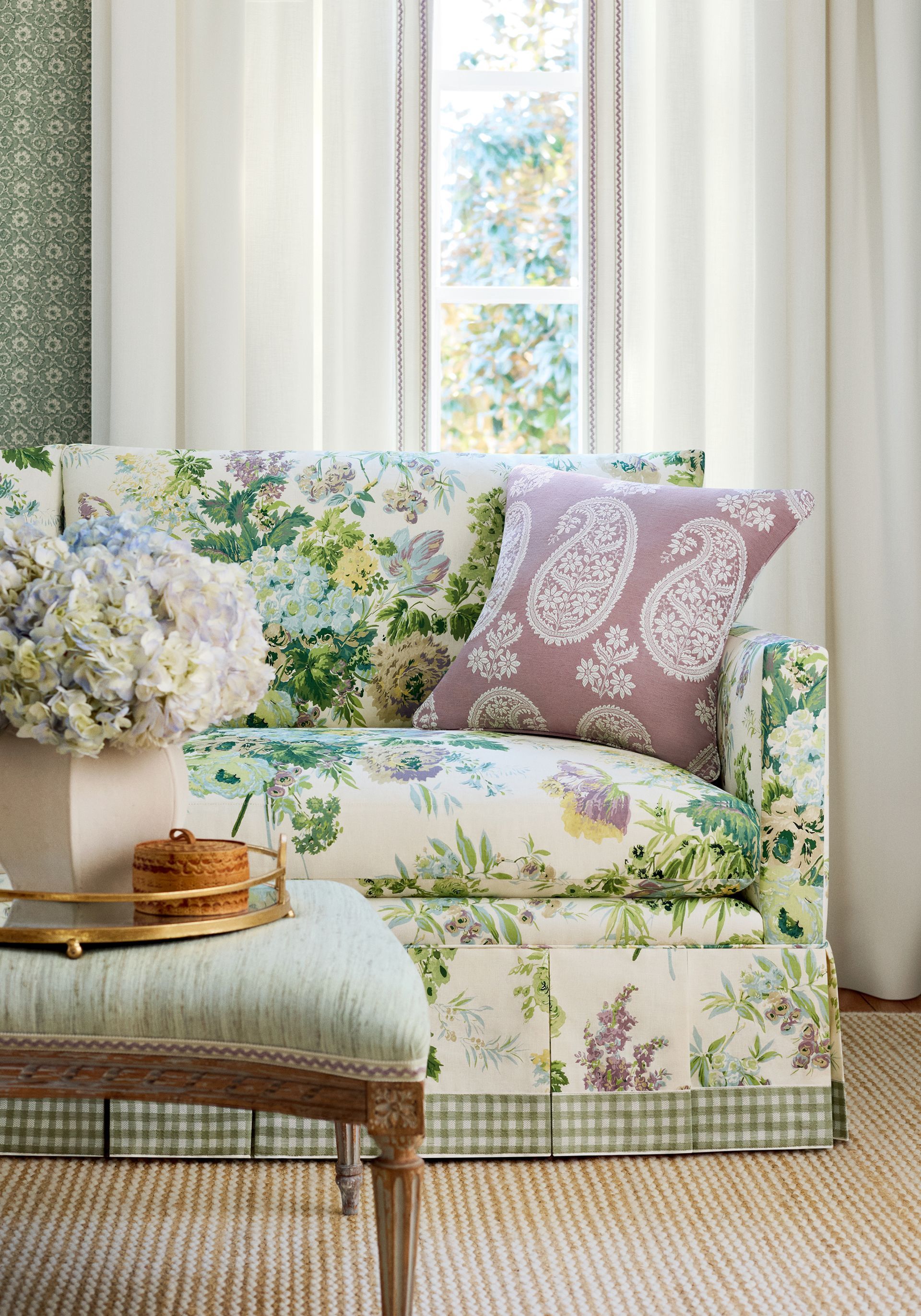 Floral print sofa with a paisley pillow, next to a window and a small stool, light-colored curtains.