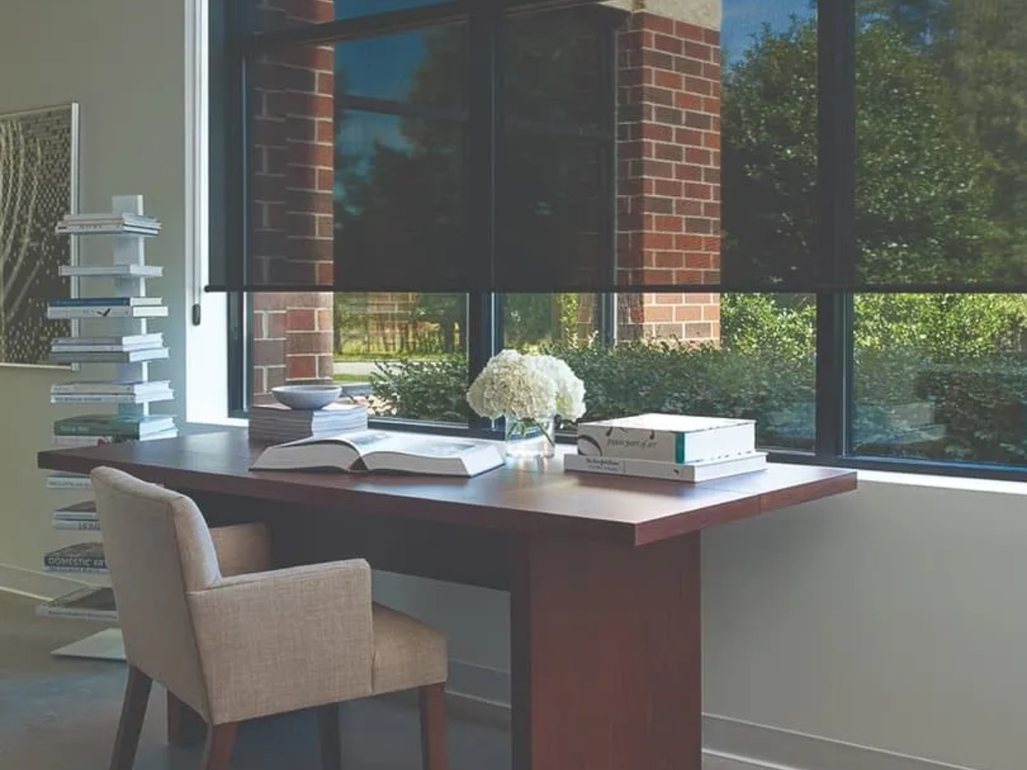 Desk with chair near a window with black shades; books, flowers, and greenery are visible.