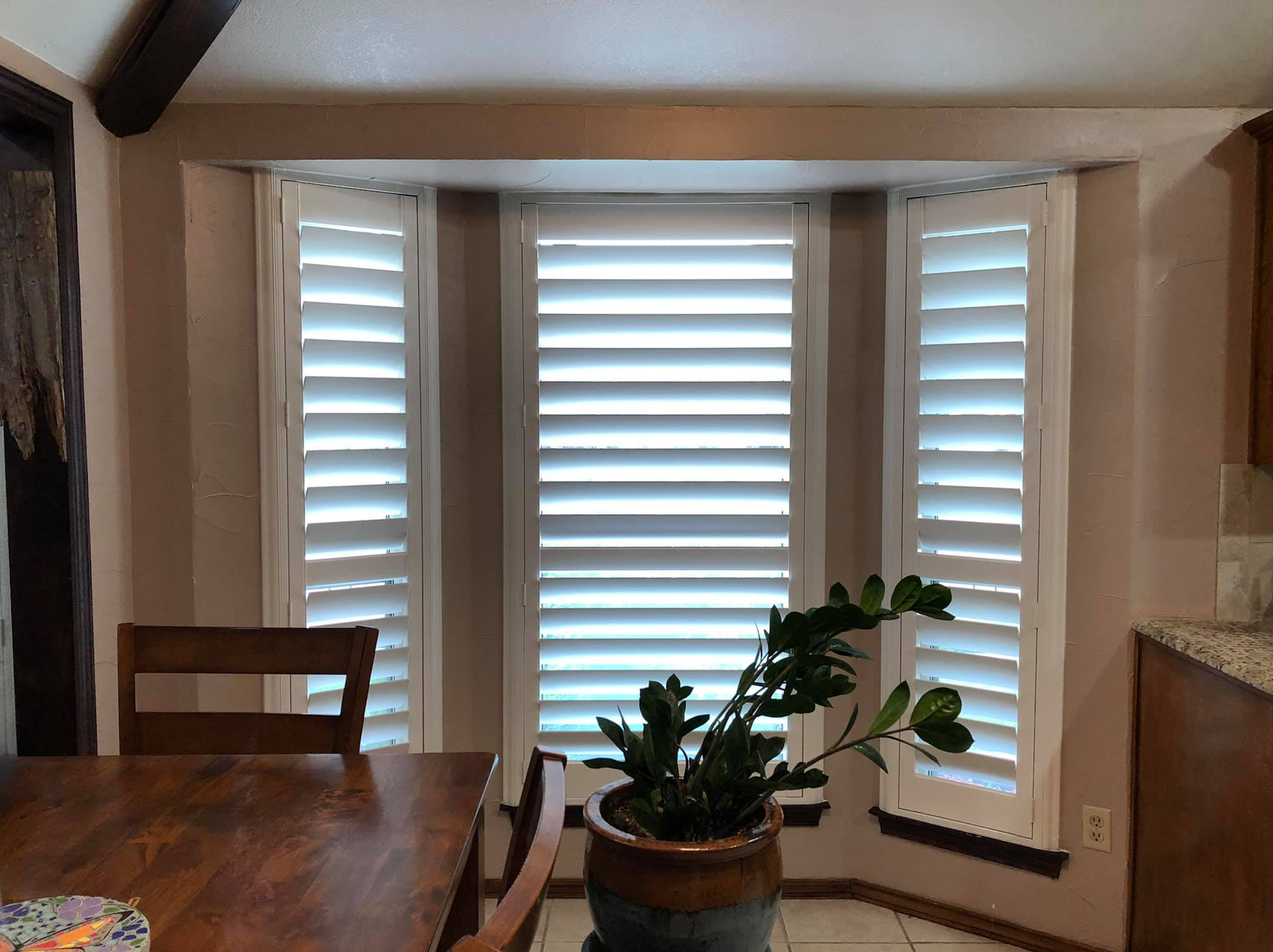 Bay window with white shutters, a potted plant, and a wooden table.