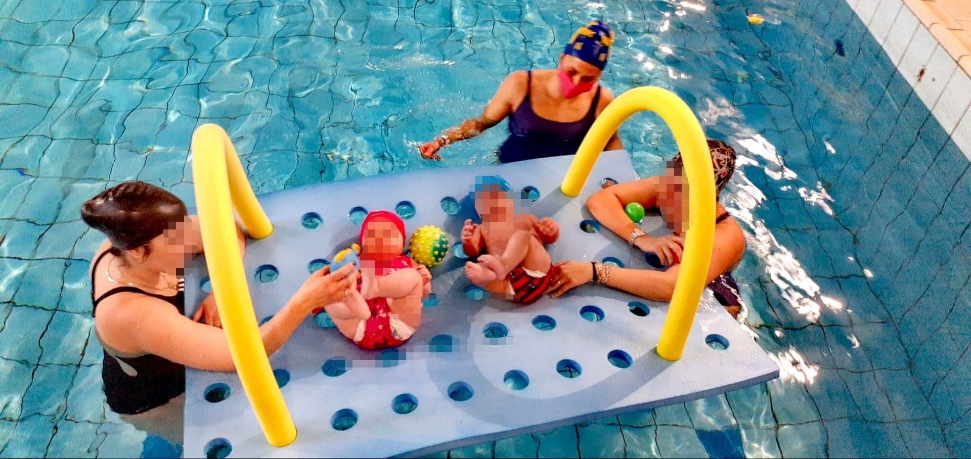 Tre neonati e tre adulti in una piscina. I neonati sono sdraiati su un materassino galleggiante blu. Gli adulti li assistono.
