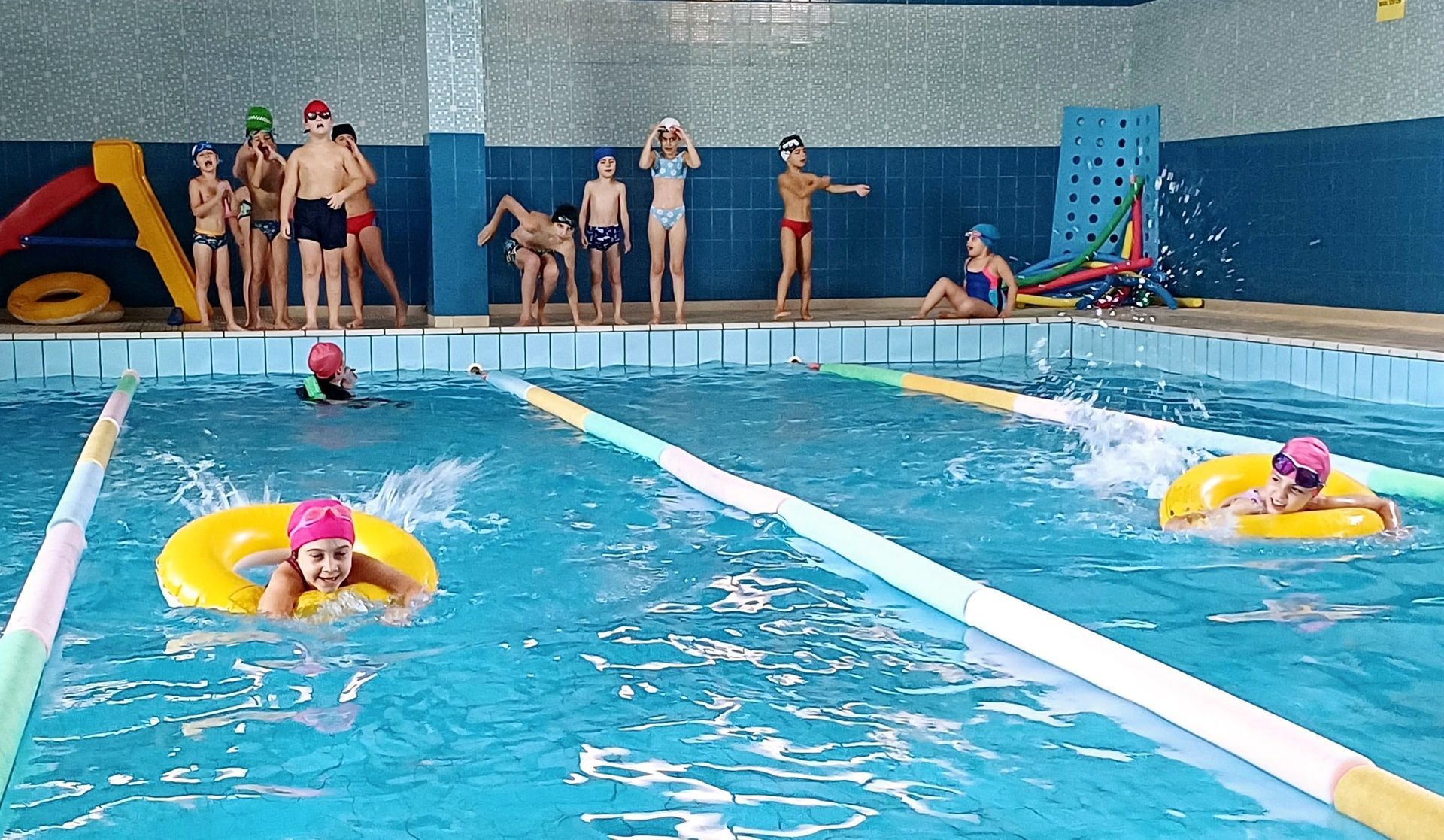 Bambini in piscina, alcuni nuotano con i galleggianti, altri stanno lì vicino. Piscina coperta con acqua azzurra e piastrelle.