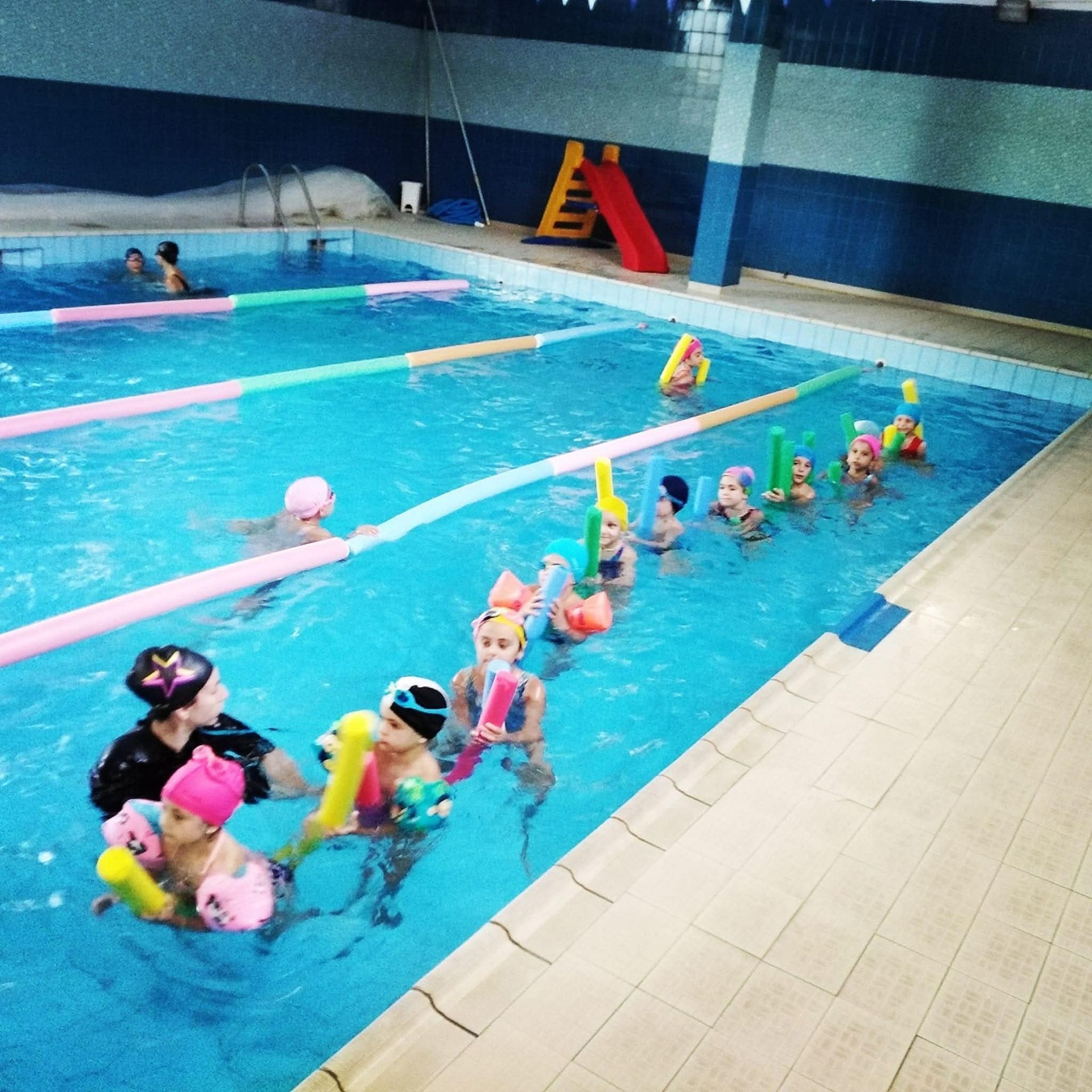 Bambini in piscina, con istruttore. I bambini indossano cuffie e usano galleggianti. Piscina coperta con pareti e acqua blu.