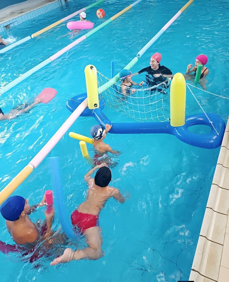 Bambini che giocano in piscina. Alcuni indossano la cuffia, uno è al centro con un cappello e un insegnante li sta istruendo.