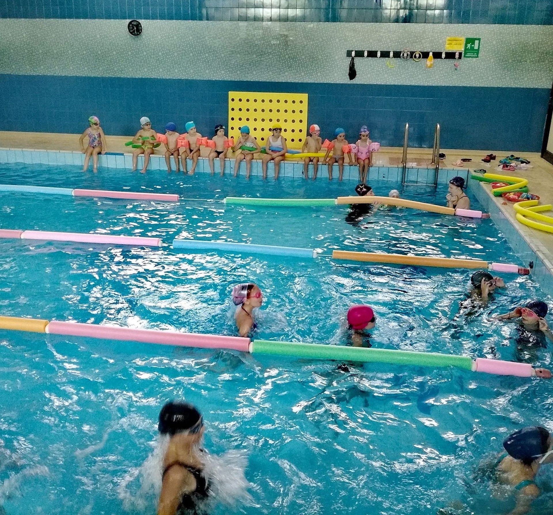 Bambini con la cuffia in piscina, che si esercitano con i galleggianti. Alcuni sono seduti sul bordo della piscina.