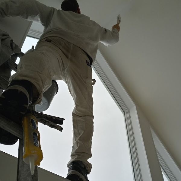 Person on a ladder painting a ceiling, wearing white overalls, indoors.