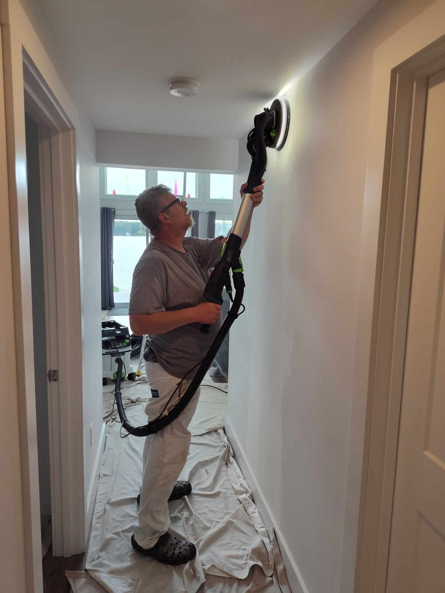 Person sanding a white wall with a long-handled sander in a hallway.