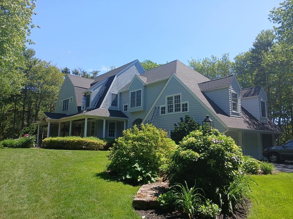 Two-story light blue house with dark roof surrounded by green trees and grass on a sunny day.