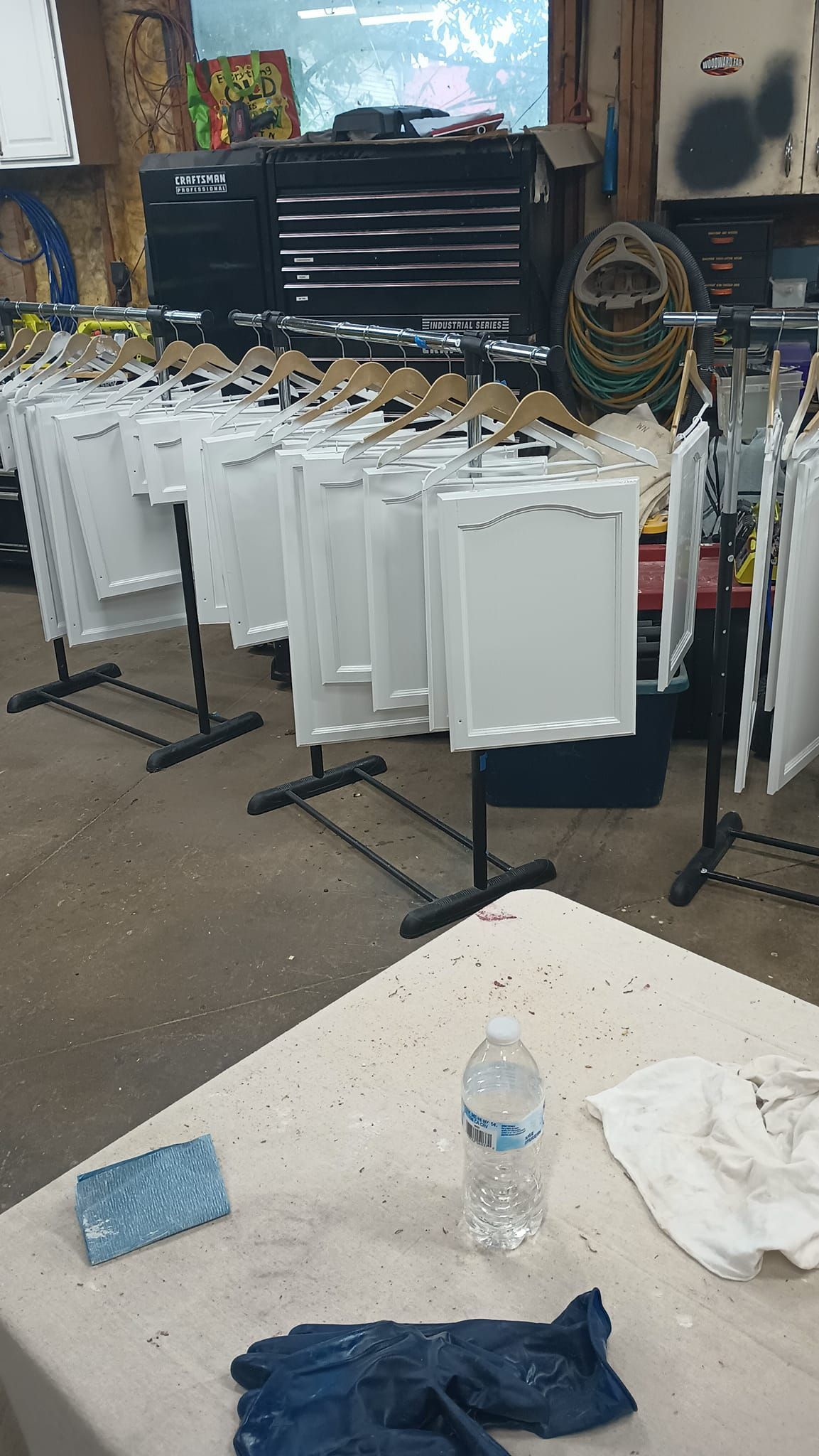 White picture frames hanging in a workshop, ready for painting. Table with water bottle and supplies in foreground.