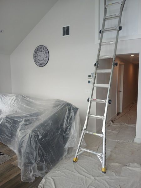 A tall ladder leans against a white wall. A covered couch, clock, and doorway are visible.