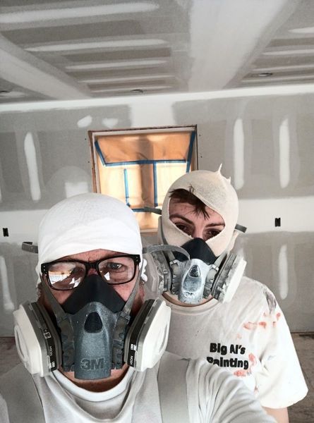 Two people wearing respirators and working in a room with drywall.