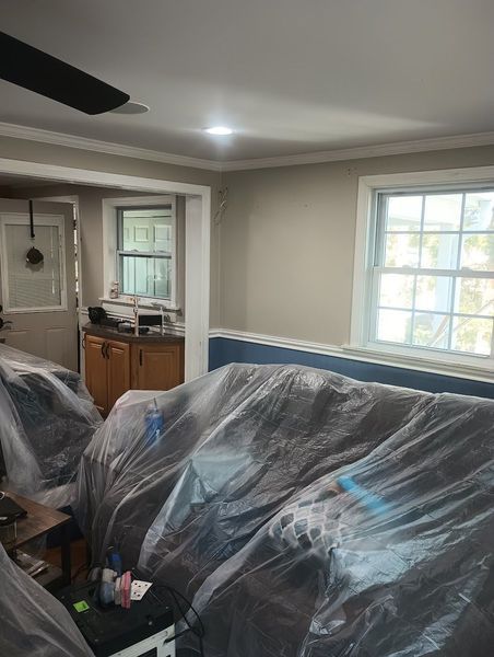 Living room with furniture covered in plastic, two windows, doorway to kitchen. Beige walls, white trim.