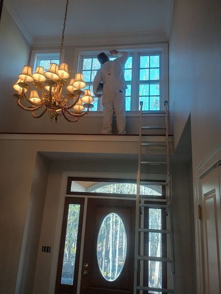 Person on platform painting window trim, using ladder, interior shot with chandelier and front door.